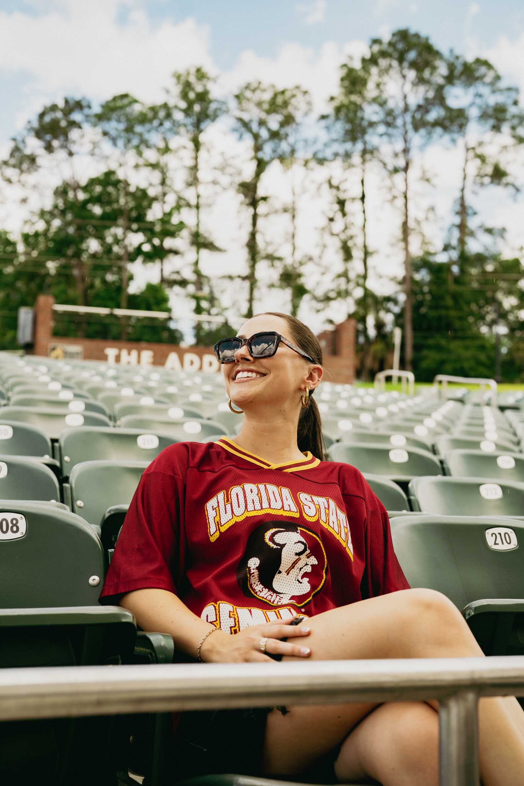 A woman wearing a Florida State Seminoles football jersey and sunglasses sitting in an outdoor stadium with empty seats and tall trees in the background.