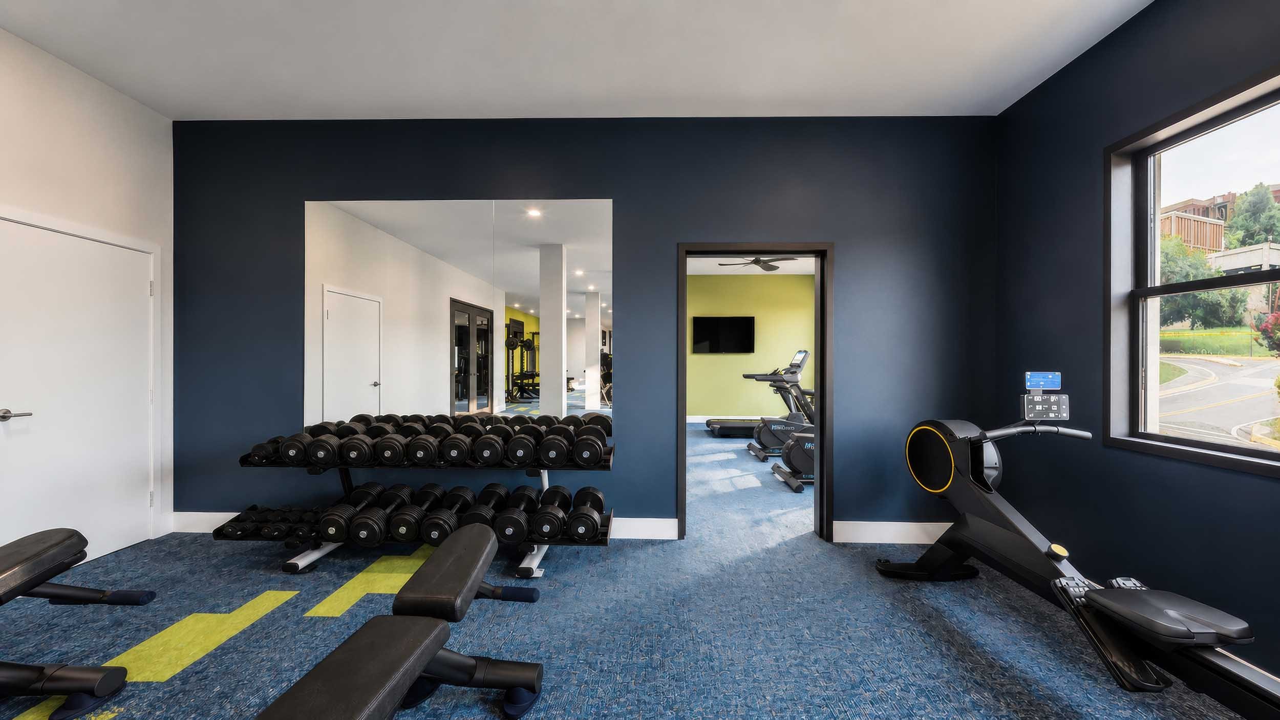 Interior of a fitness room with dumbbells, workout benches, a treadmill, and exercise bikes, with a window showing outdoor scenery.