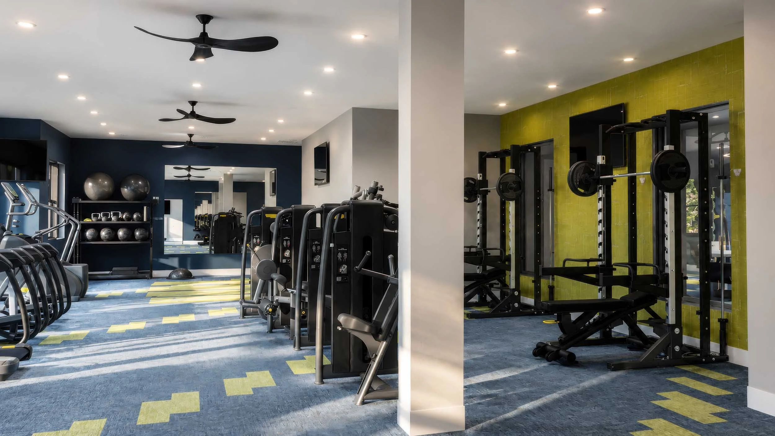 Fitness center with cardio machines, free weights, and weightlifting equipment, featuring a colorful floor and ceiling fans.