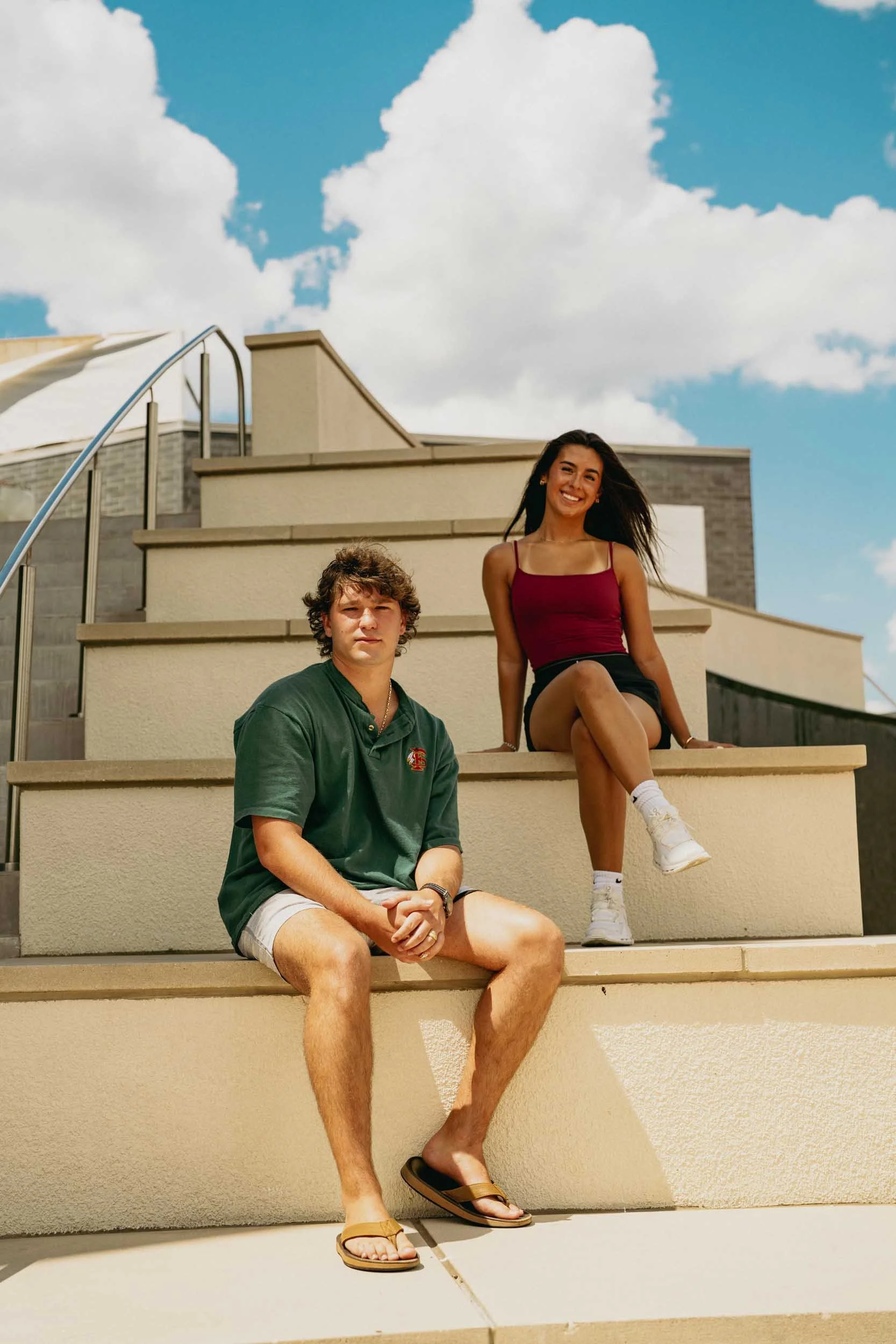 Two young people sitting on outdoor stairs under a partly cloudy sky. The person in front is a young man with curly hair, wearing a green shirt, shorts, and flip-flops. The person behind is a young woman with long dark hair, wearing a red tank top, black shorts, white socks, and white sneakers. She is smiling and has one raised knee.