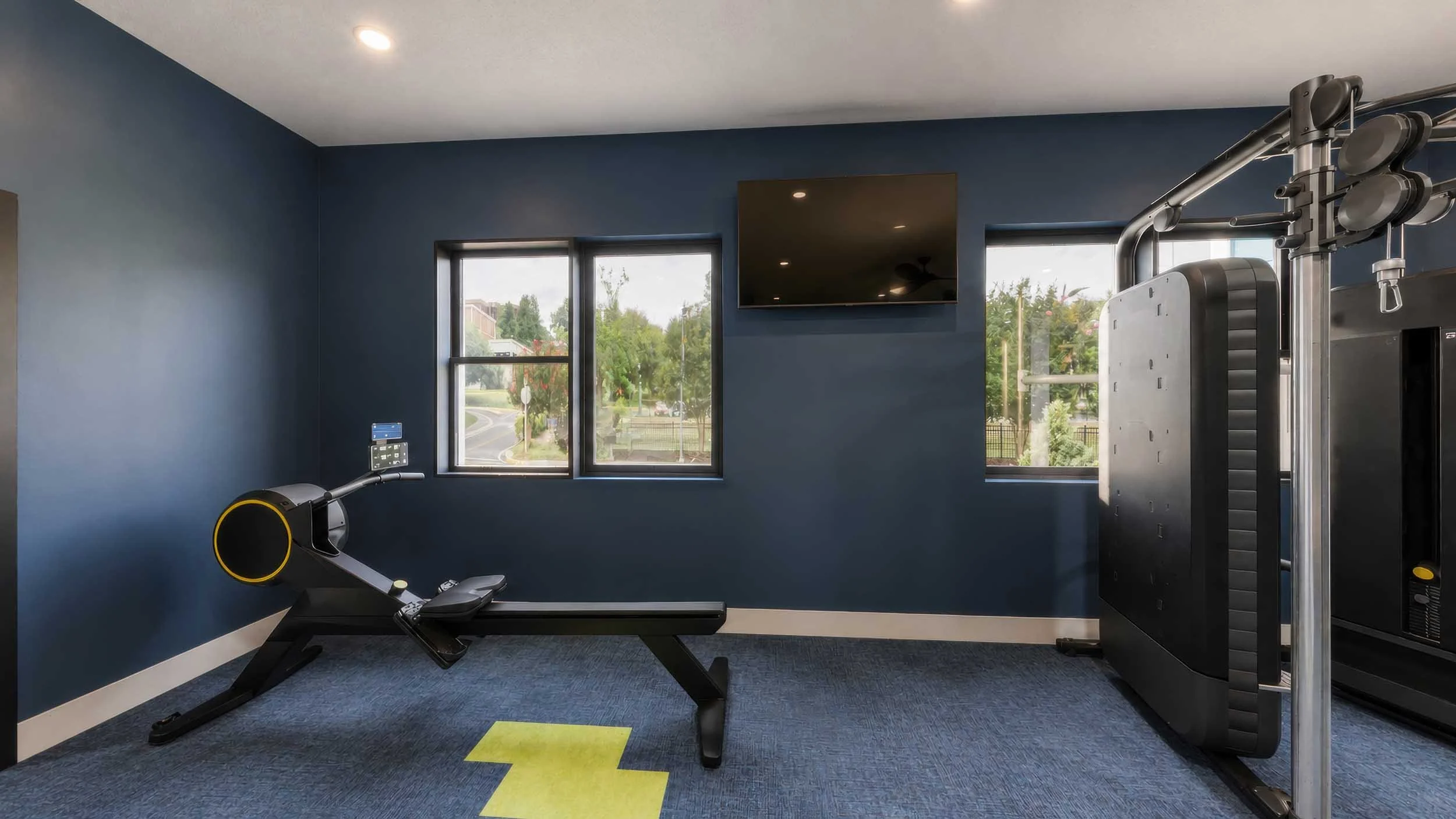 Home gym with a treadmill, various exercise equipment, and large windows showing a view of trees and buildings outside.