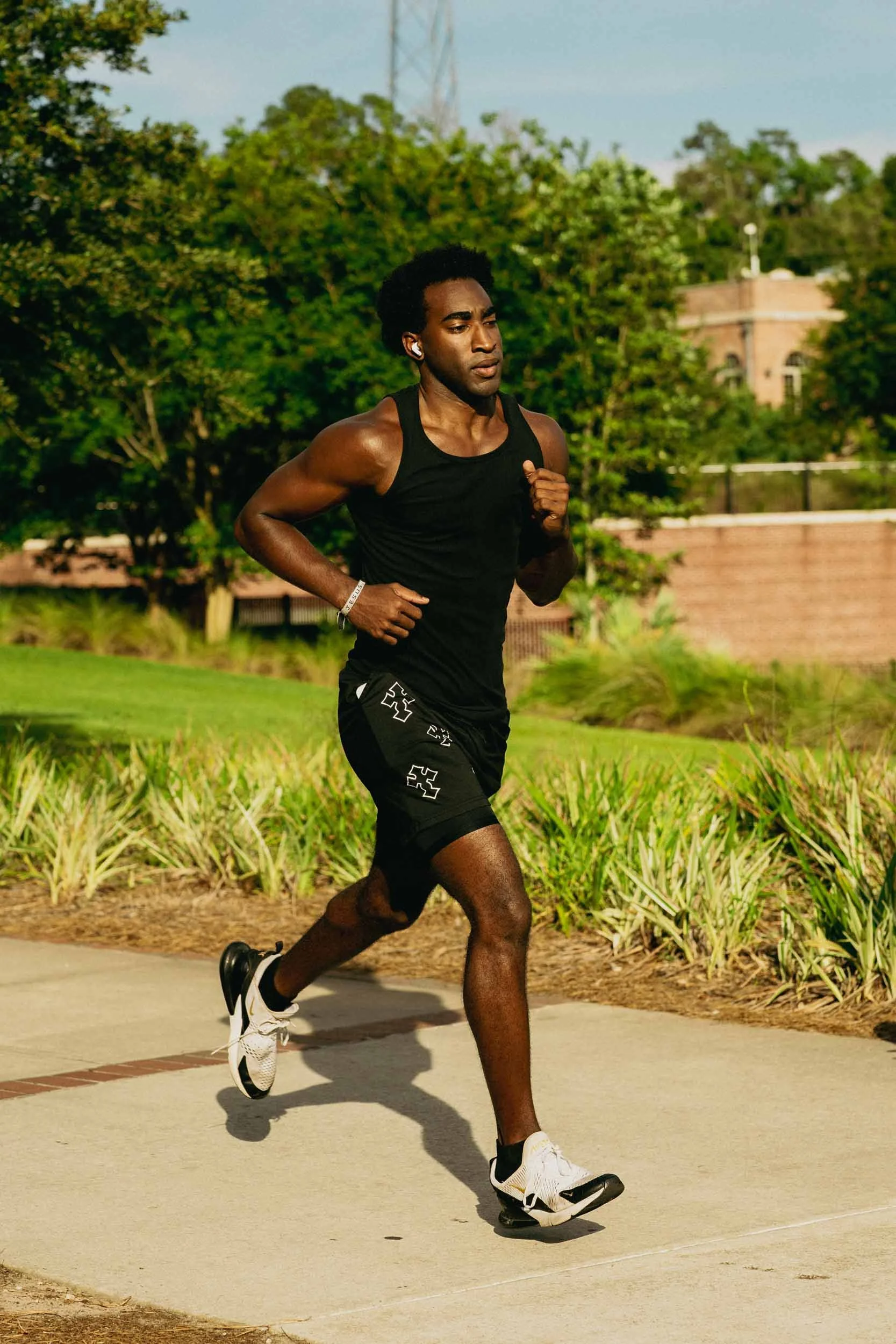 A young man runs outdoors on a sunny day, dressed in athletic clothing, wearing earbuds, with lush green trees and a building in the background.