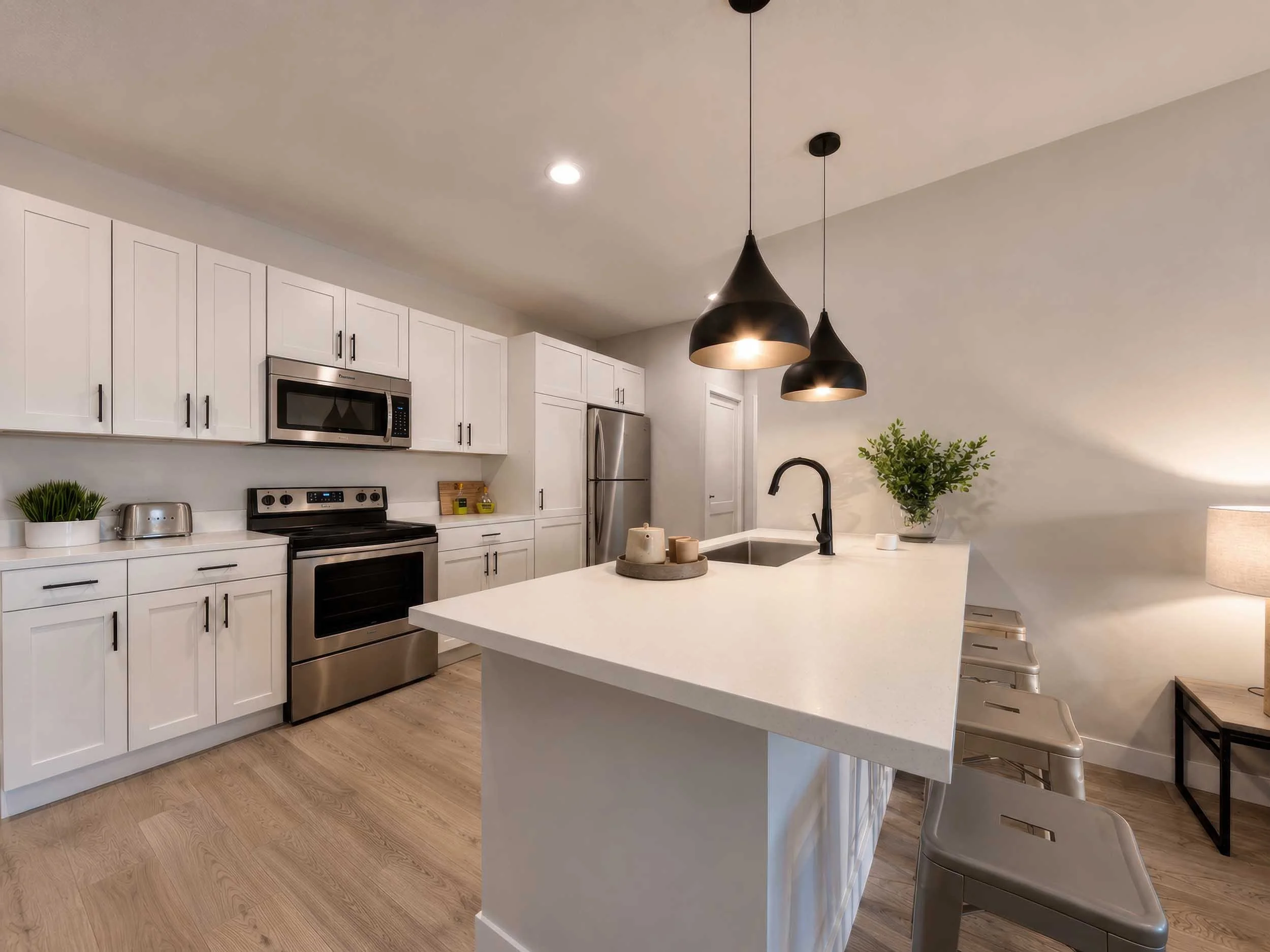 Modern kitchen with white cabinets, stainless steel appliances, black pendant lights, a kitchen island with bar stools, potted plant, and light wood flooring.