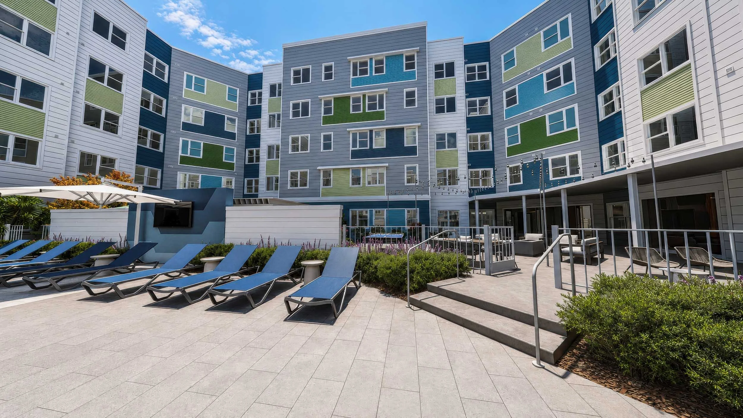 Outdoor pool area with lounge chairs and an umbrella in front of a colorful multi-story apartment complex under a partly cloudy sky.