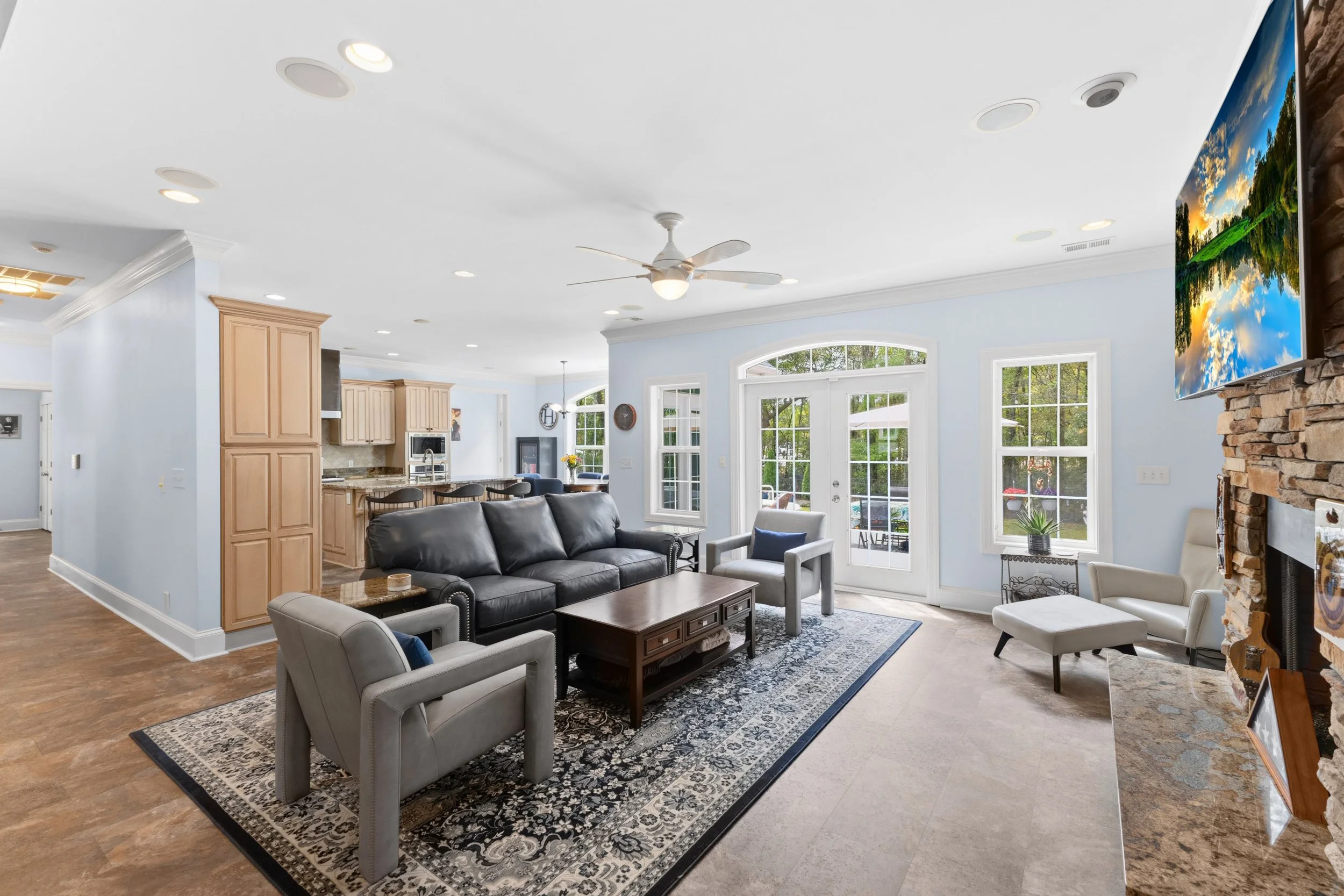 Bright living room with light blue walls, large windows, a stone fireplace with a mounted flat-screen TV, black leather sofa, light gray armchairs, a wooden coffee table, and a patterned rug. Open kitchen with wooden cabinets and a breakfast bar in t