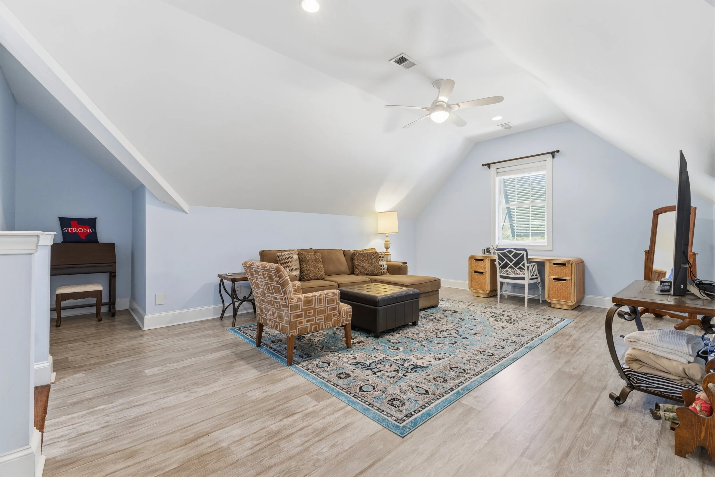 A spacious living room with light blue walls, a brown sectional sofa, a patterned armchair, and a black ottoman on a decorative area rug. There is a window with white trim, a framed mirror, a desk with a chair, and a small table with a lamp. The room