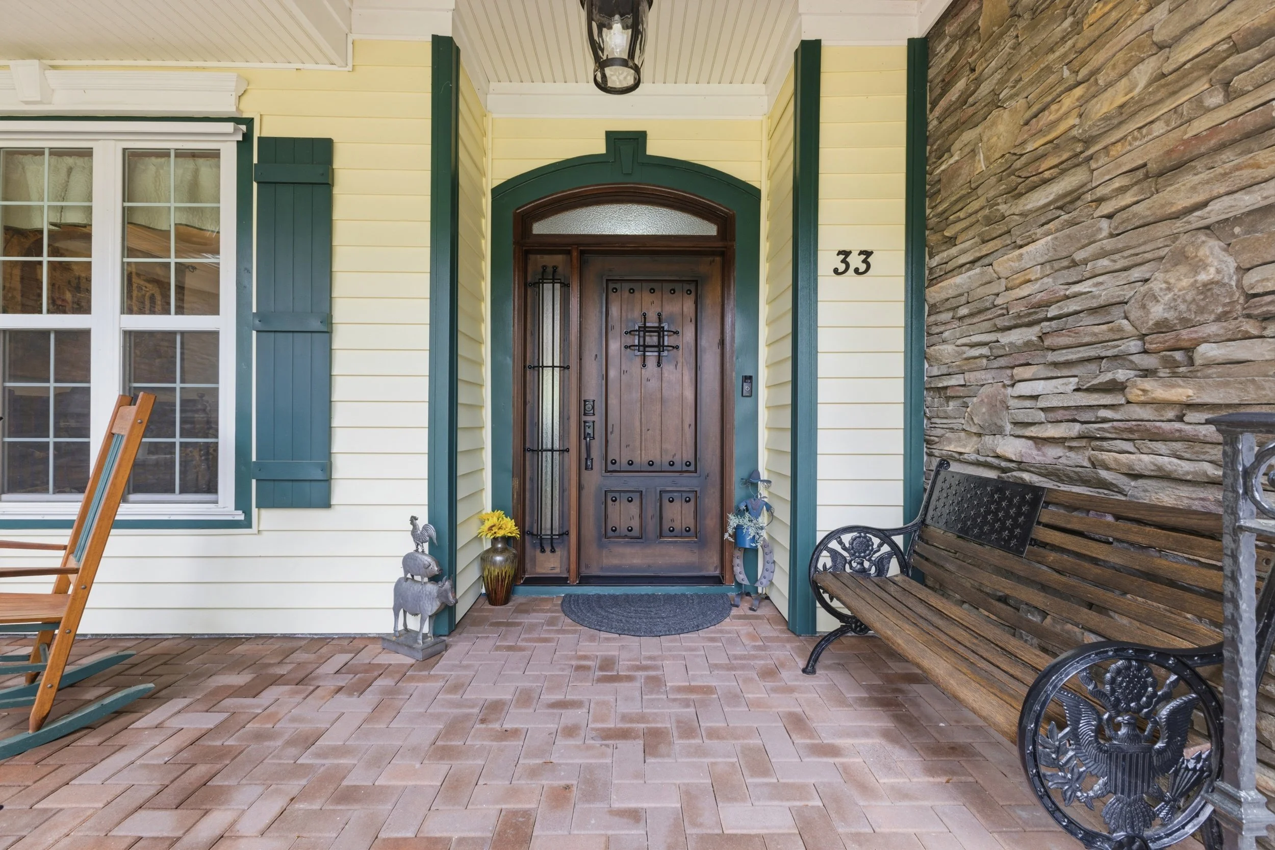 Front porch of a house with a dark wooden front door, a black metal bench on the right, a rocking chair on the left, a window with green shutters, and decorative items including a yellow flower vase, a dog figurine, and a horseshoe.