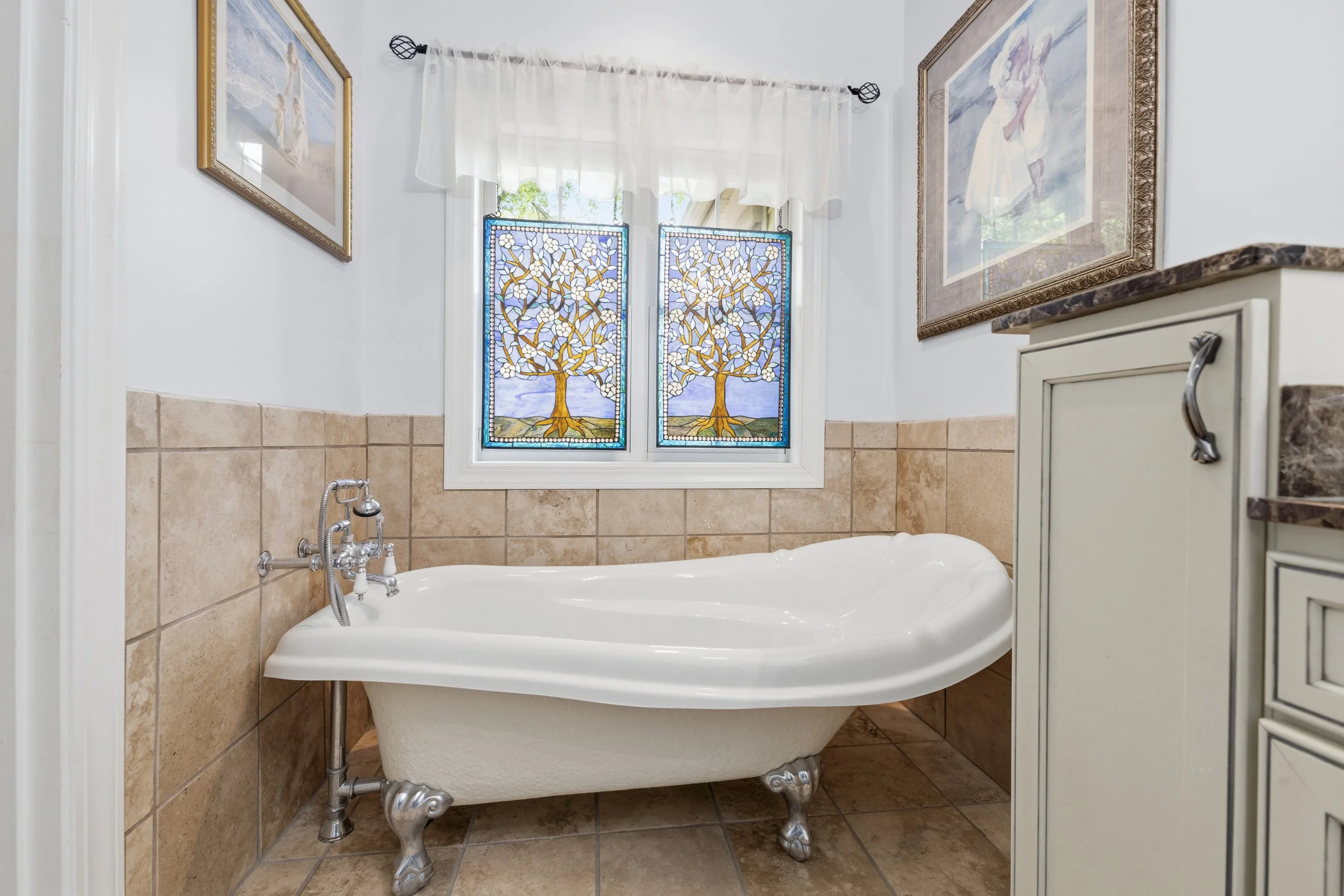 A vintage-style clawfoot bathtub in a bathroom, with stained glass windows featuring a tree design and framed artwork on the walls.