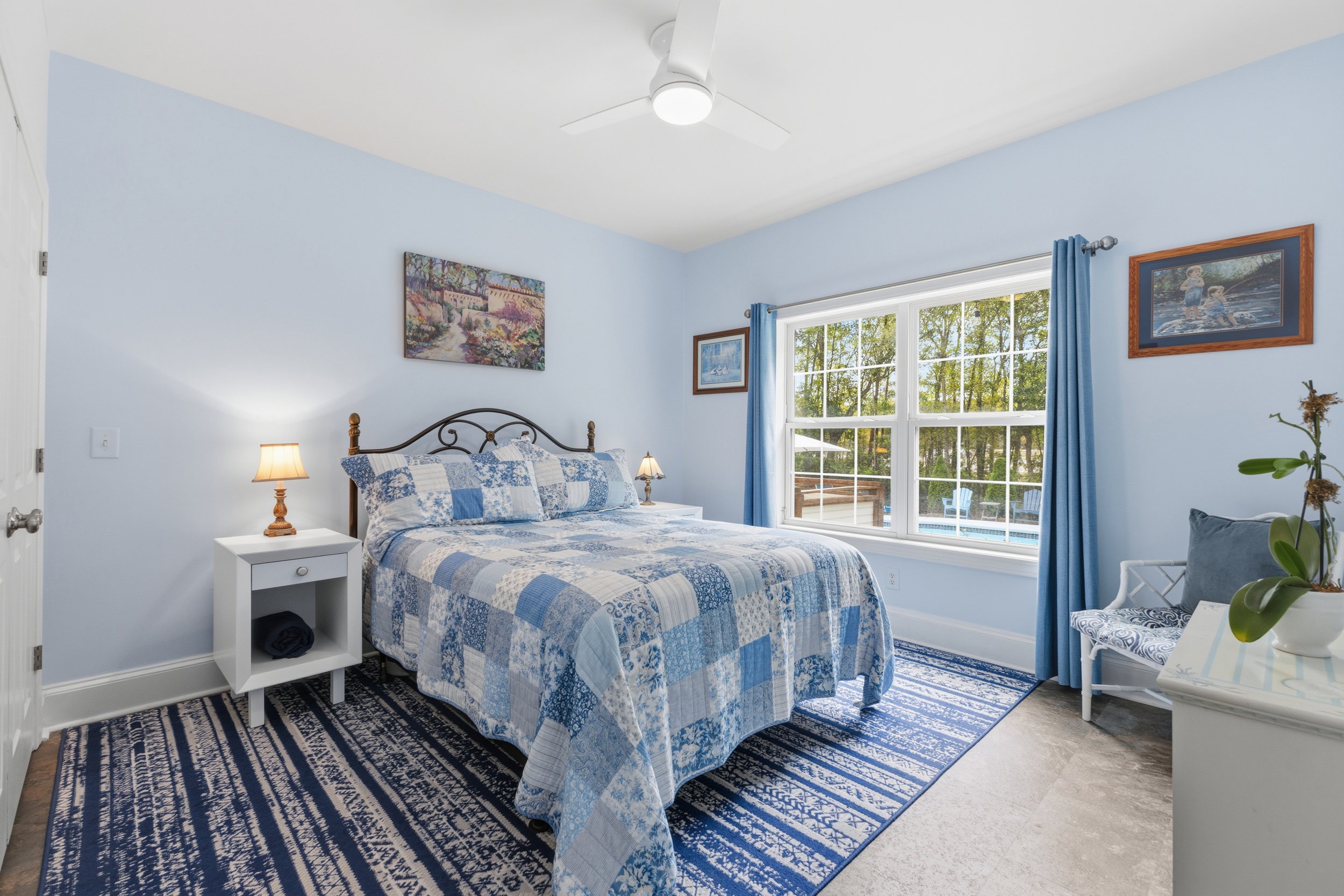 Bedroom with light blue walls, a bed with a blue and white patchwork quilt, two bedside lamps, a white bedside table, a large window with blue curtains, a blue patterned rug, a white chair with a gray pillow, and framed artwork on the walls.