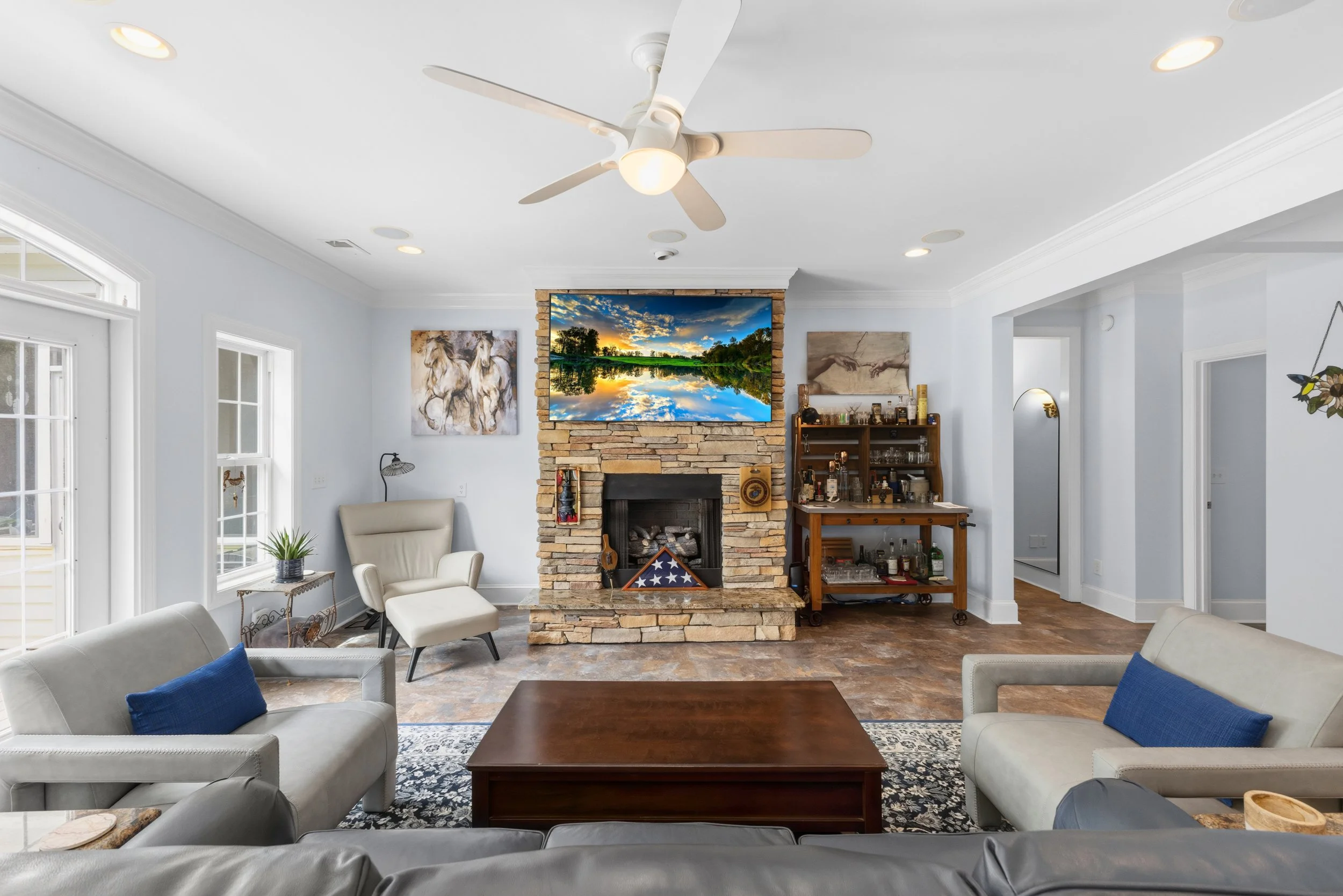 Living room with white walls, ceiling fan, and a stone fireplace with a large flat-screen TV above it. There are cream and gray sofas, a wooden coffee table, an armchair, and a bar cart. Decor includes paintings and a plant, with hardwood floors and 