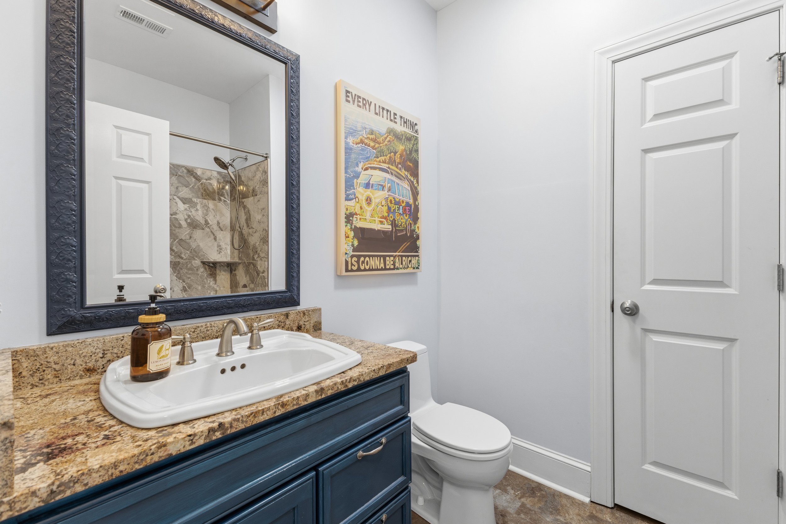 A bathroom with a blue vanity, granite countertop, white sink, and a brown soap dispenser. A large blue-framed mirror hangs above the vanity. To the right, there is a white toilet next to a wall with a colorful poster of a van and the text "Every lit