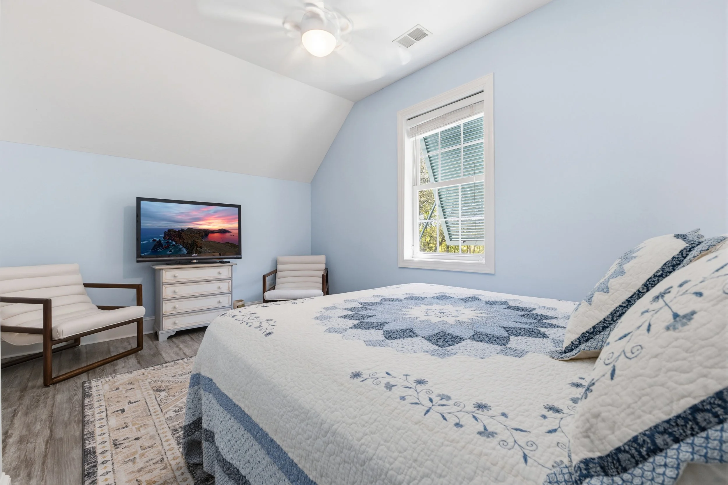 A cozy bedroom with light blue walls, a white quilt with blue patterns on a bed, a window with white trim and blinds, a ceiling fan with light, a TV on top of a small white dresser, two white chairs with wooden frames, and a patterned rug.