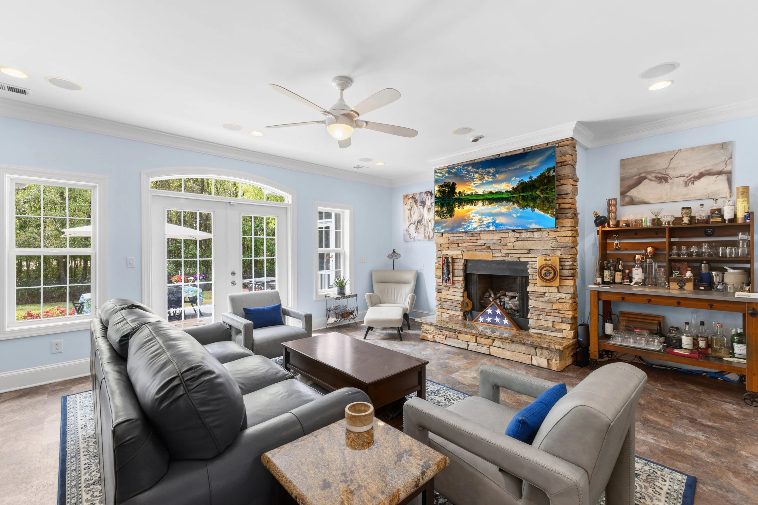 Living room with light blue walls, a stone fireplace with a mounted flat-screen TV, large windows and door leading outside, gray sofa, white armchairs, and a wooden bar cart.