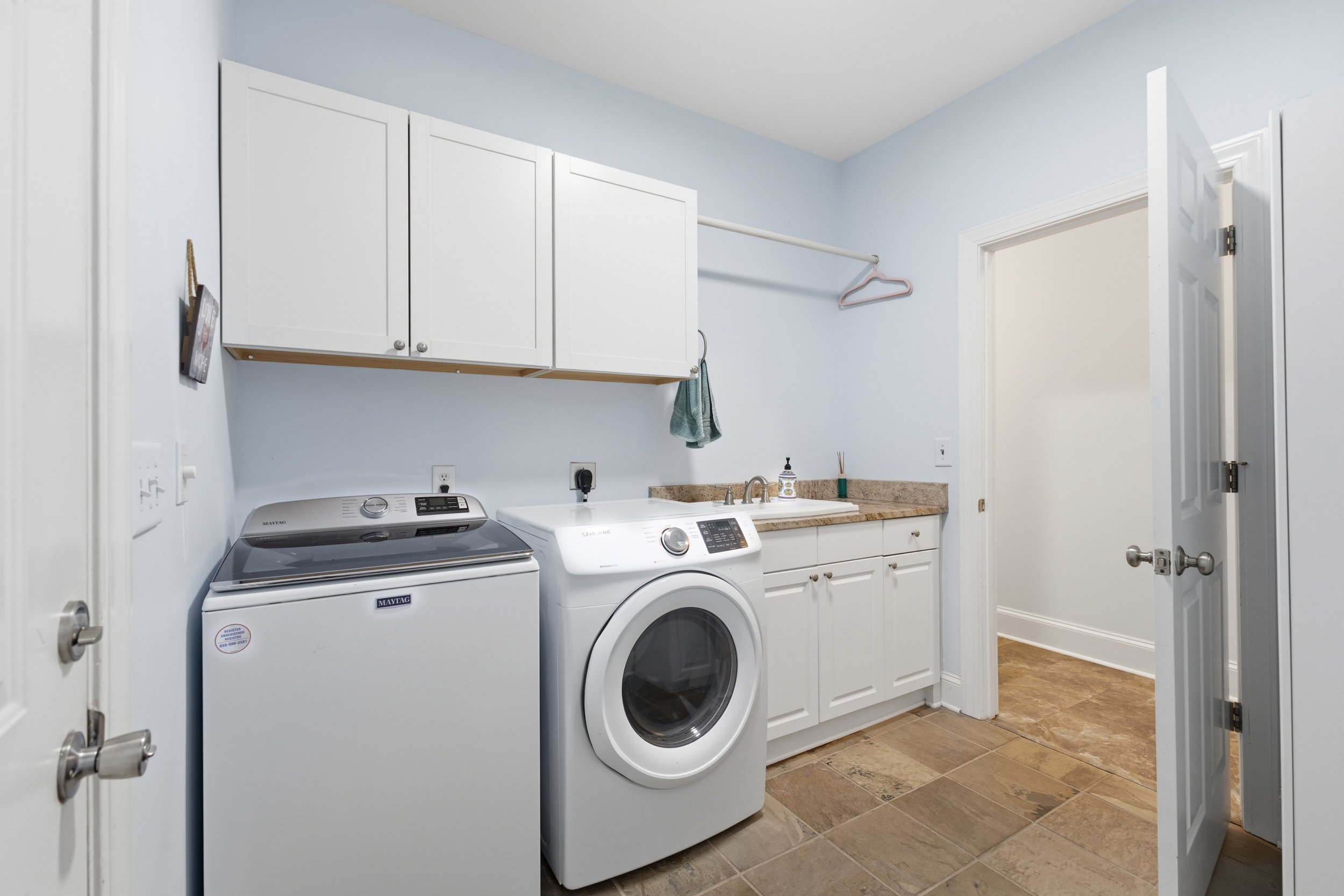 Laundry room with white cabinets, a washer and dryer, a sink, and a countertop, with light blue walls and brown tiled floor.