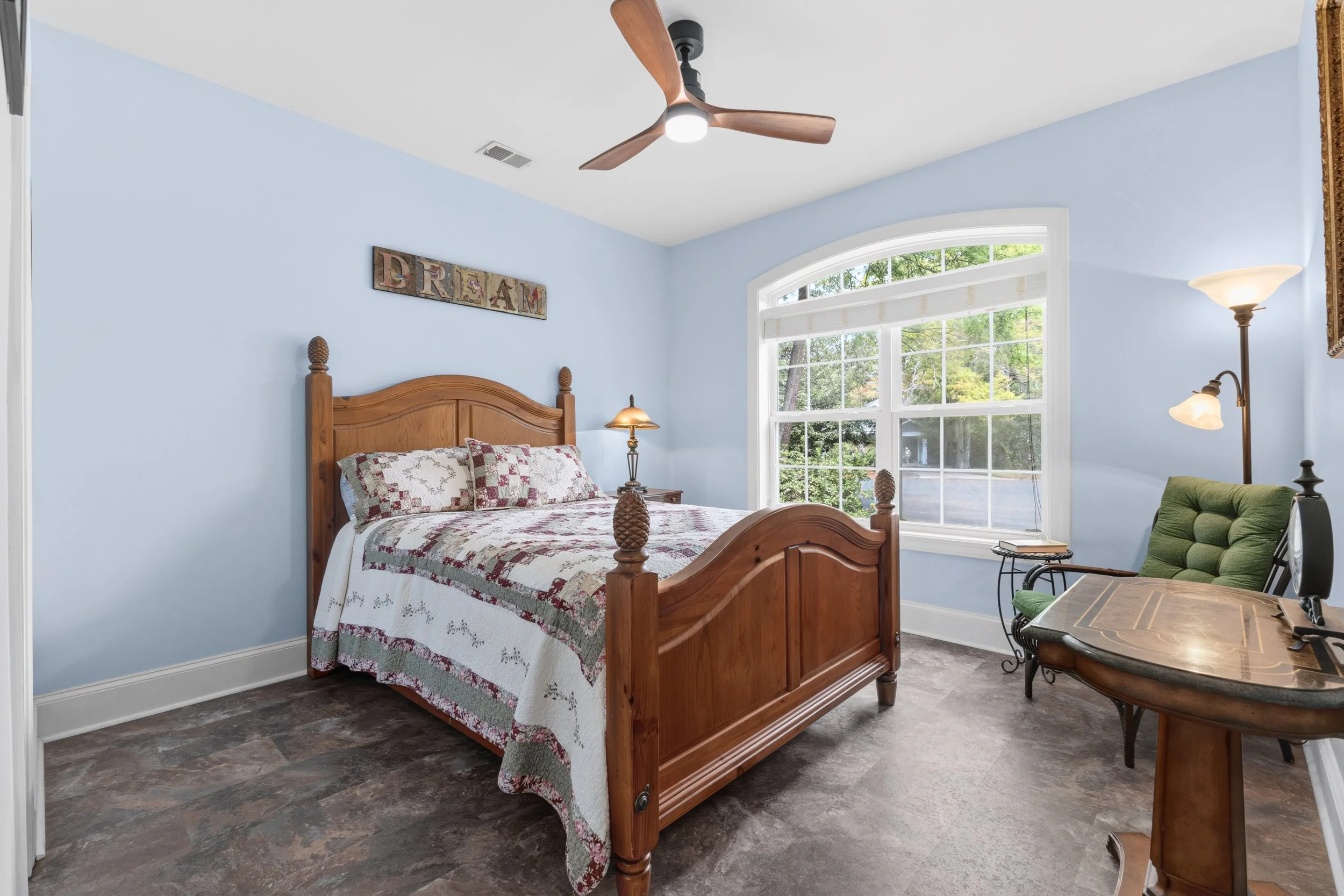 A cozy bedroom with a wooden bed frame, quilted bedding, and decorative pillows. Sunlight streams in through a large window, illuminating a reading chair and a small desk, with a wall-mounted 'DREAM' sign above the bed.