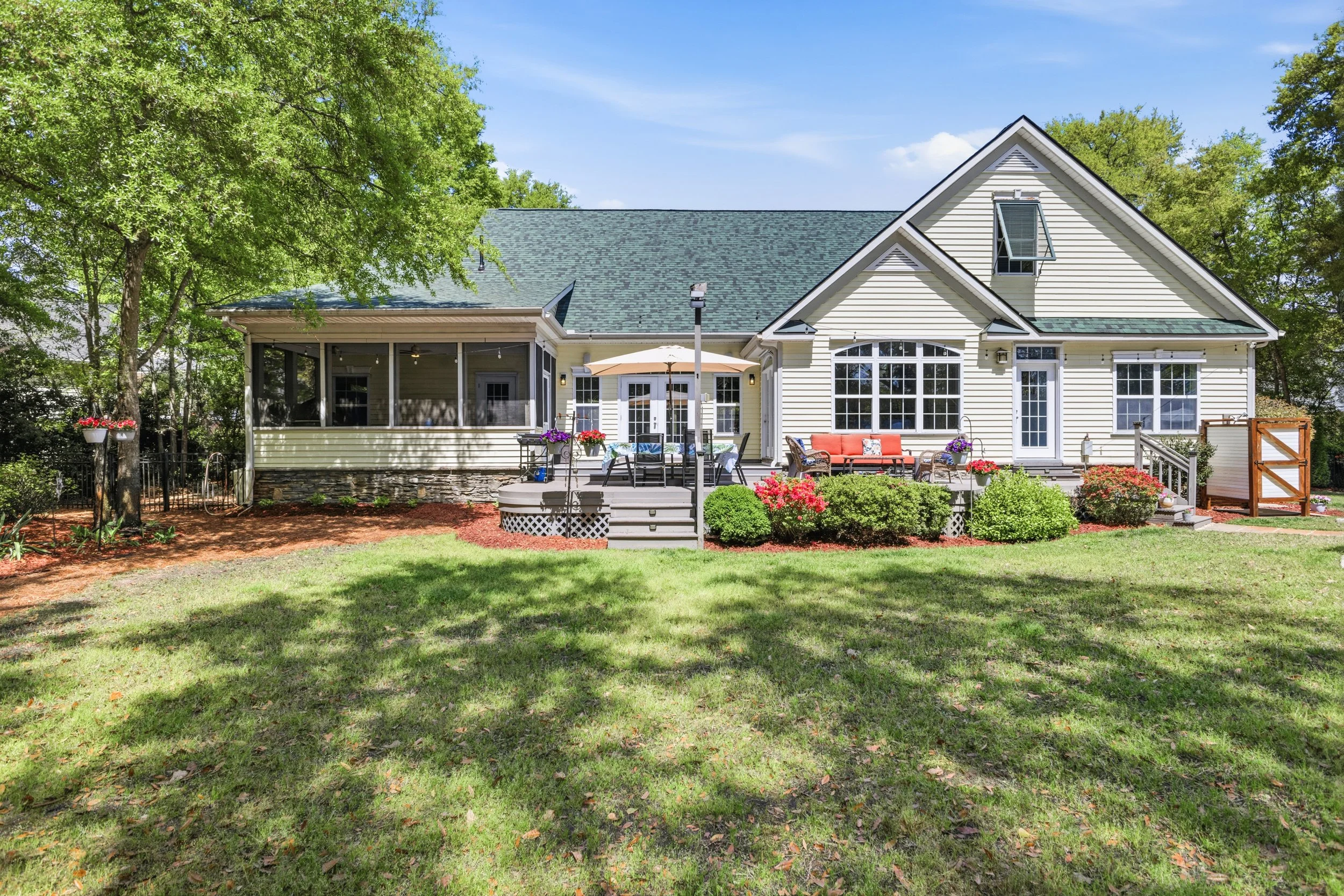 A backyard with a well-maintained lawn, trees, and a house with a screened porch and a deck. The deck has outdoor furniture, potted plants, and an umbrella, while the house features large windows and a steep roof.