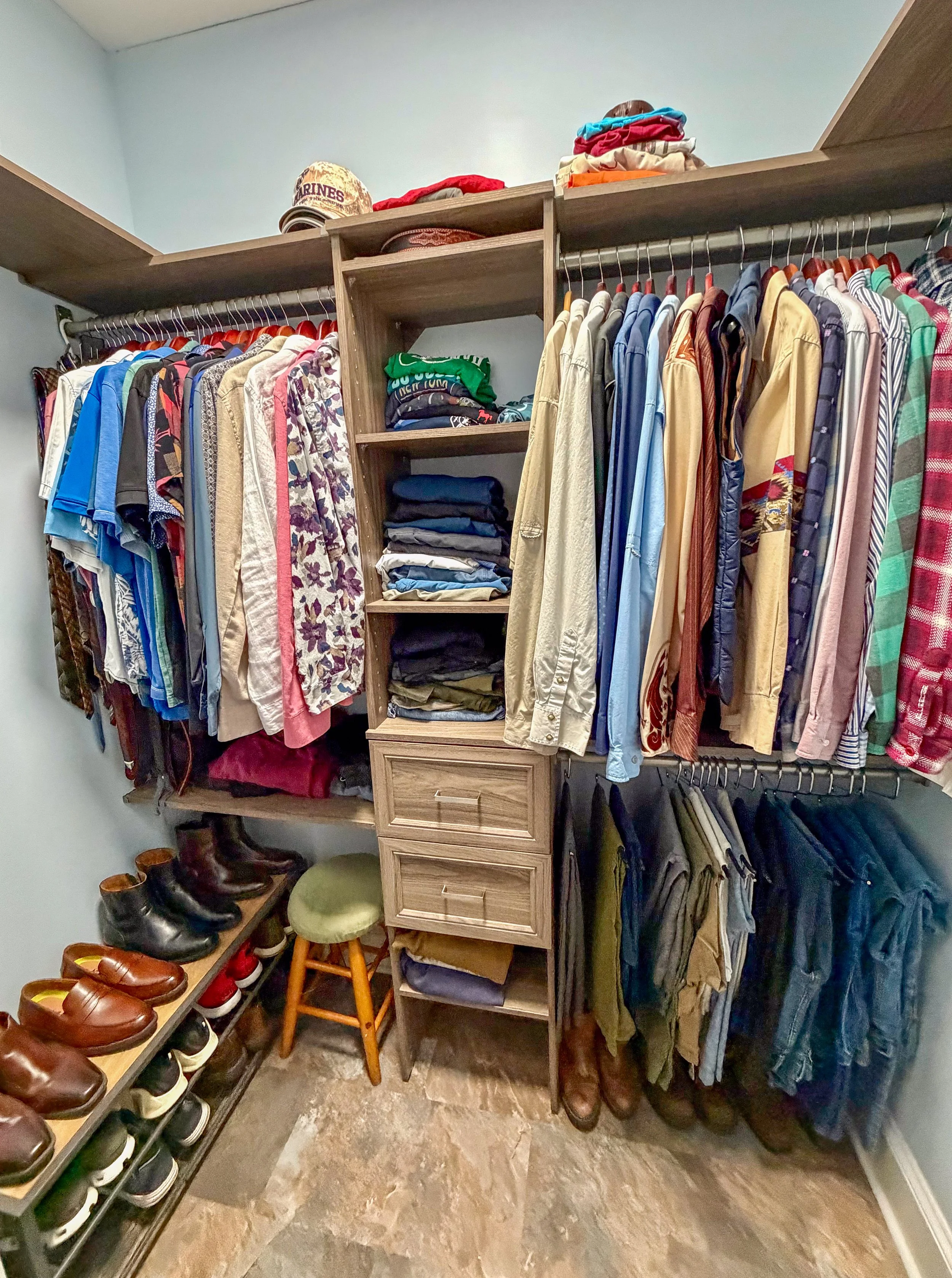A walk-in closet with shelves and hanging rods filled with clothing, shoes on the floor, and a small green stool in the corner.