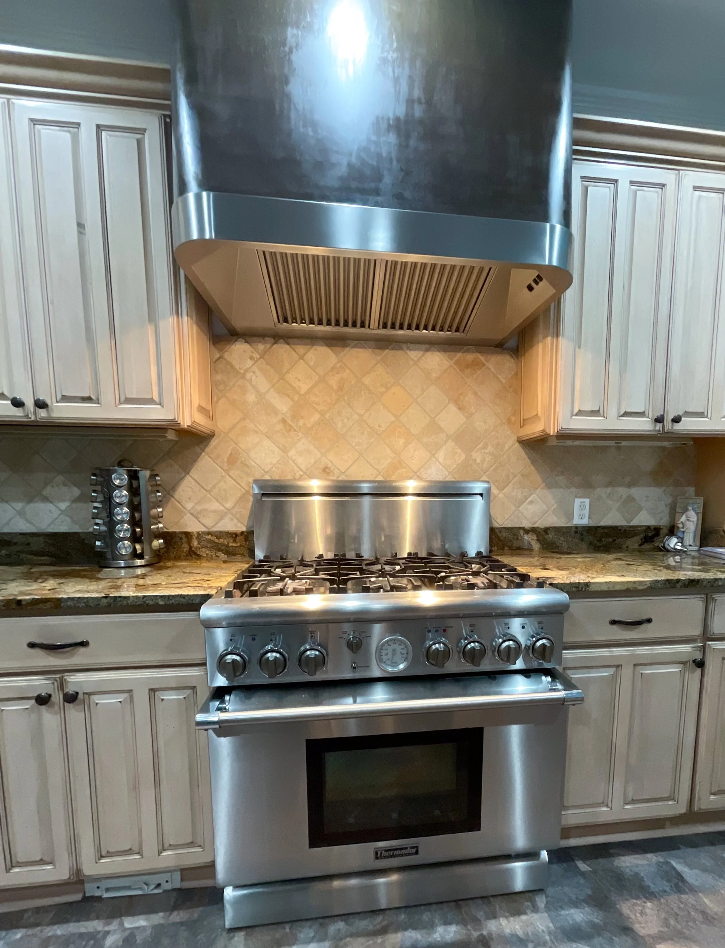 Stainless steel gas stove with oven, beige granite countertops, cream cabinet drawers, tiled backsplash, and a stainless steel range hood in a kitchen.