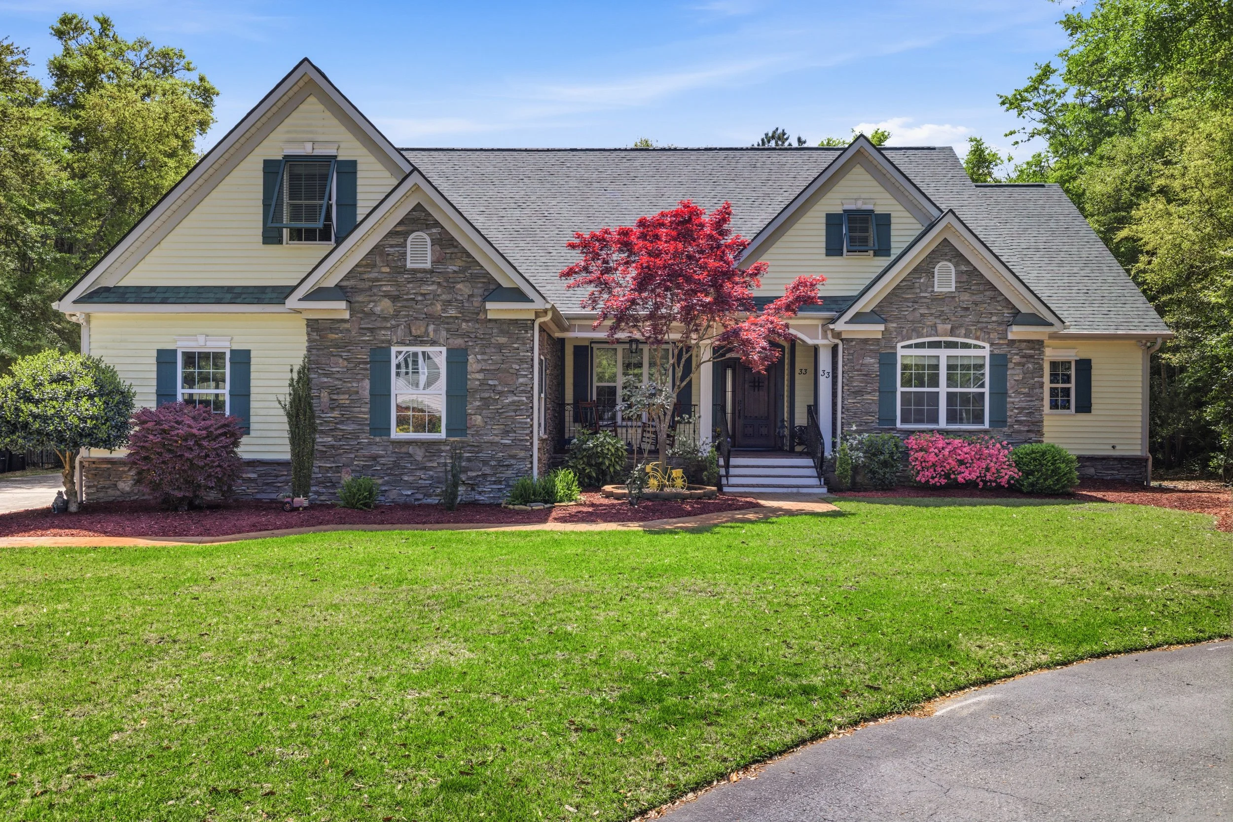 A two-story house with a stone and yellow siding exterior, surrounded by green grass and landscaped plants, with a tree in front and a driveway on the side.