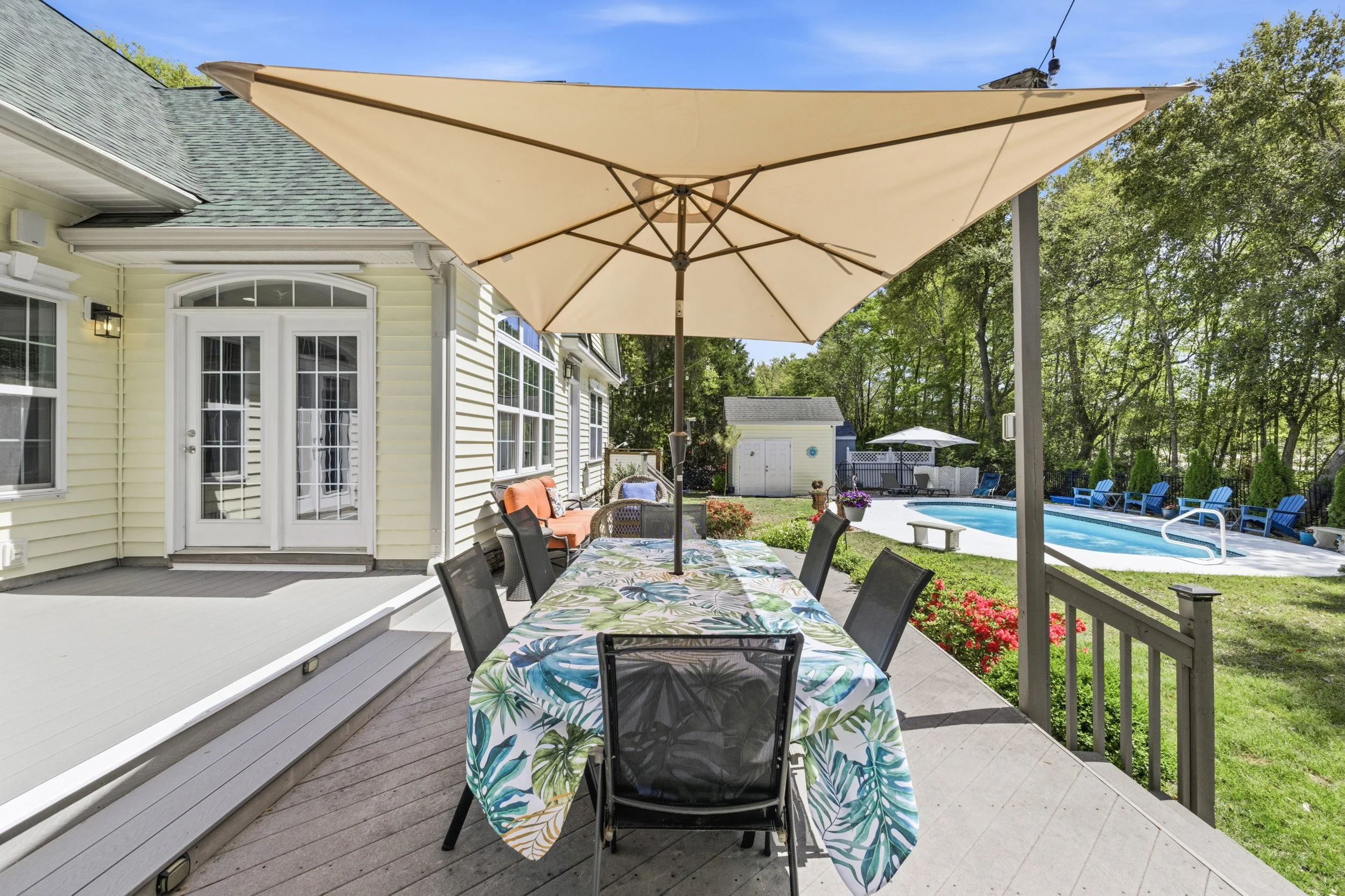 Backyard patio with an outdoor dining table, eight chairs, beige umbrella, pool with lounge chairs, small shed, green trees, and garden.