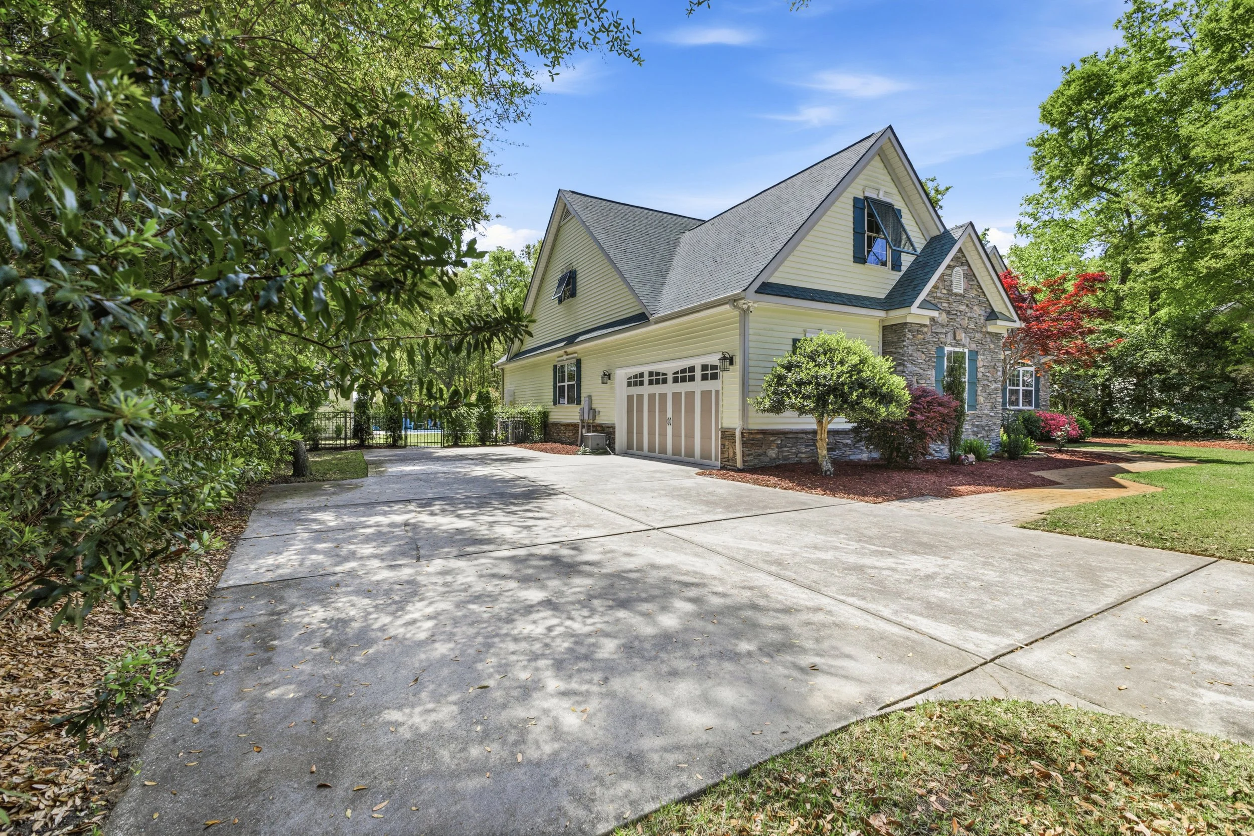 A two-story house with a stone and siding exterior, a gray roof, and a spacious driveway surrounded by greenery and trees.
