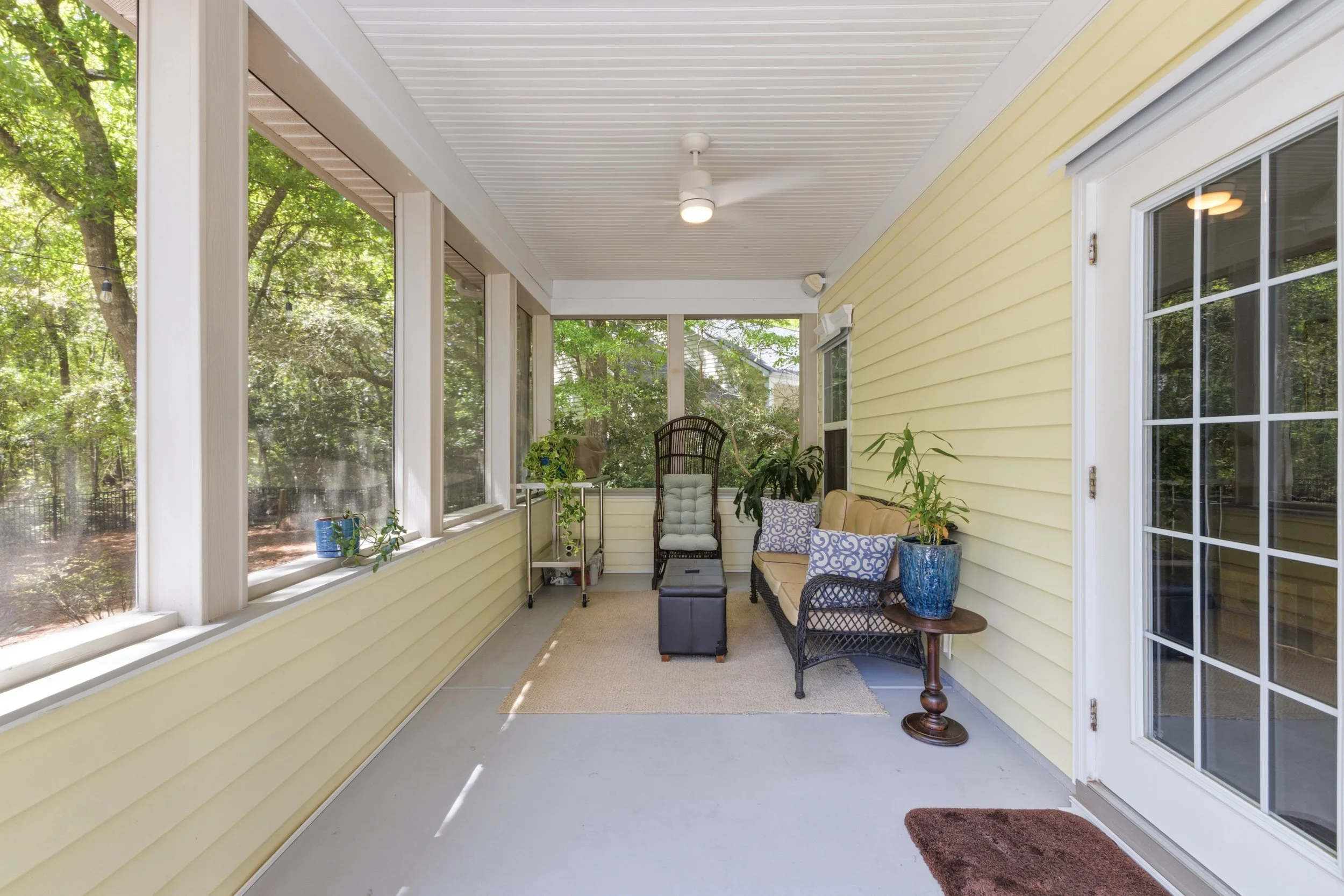 Screened porch with yellow siding, glass doors, ceiling fan, and outdoor seating, surrounded by green trees.