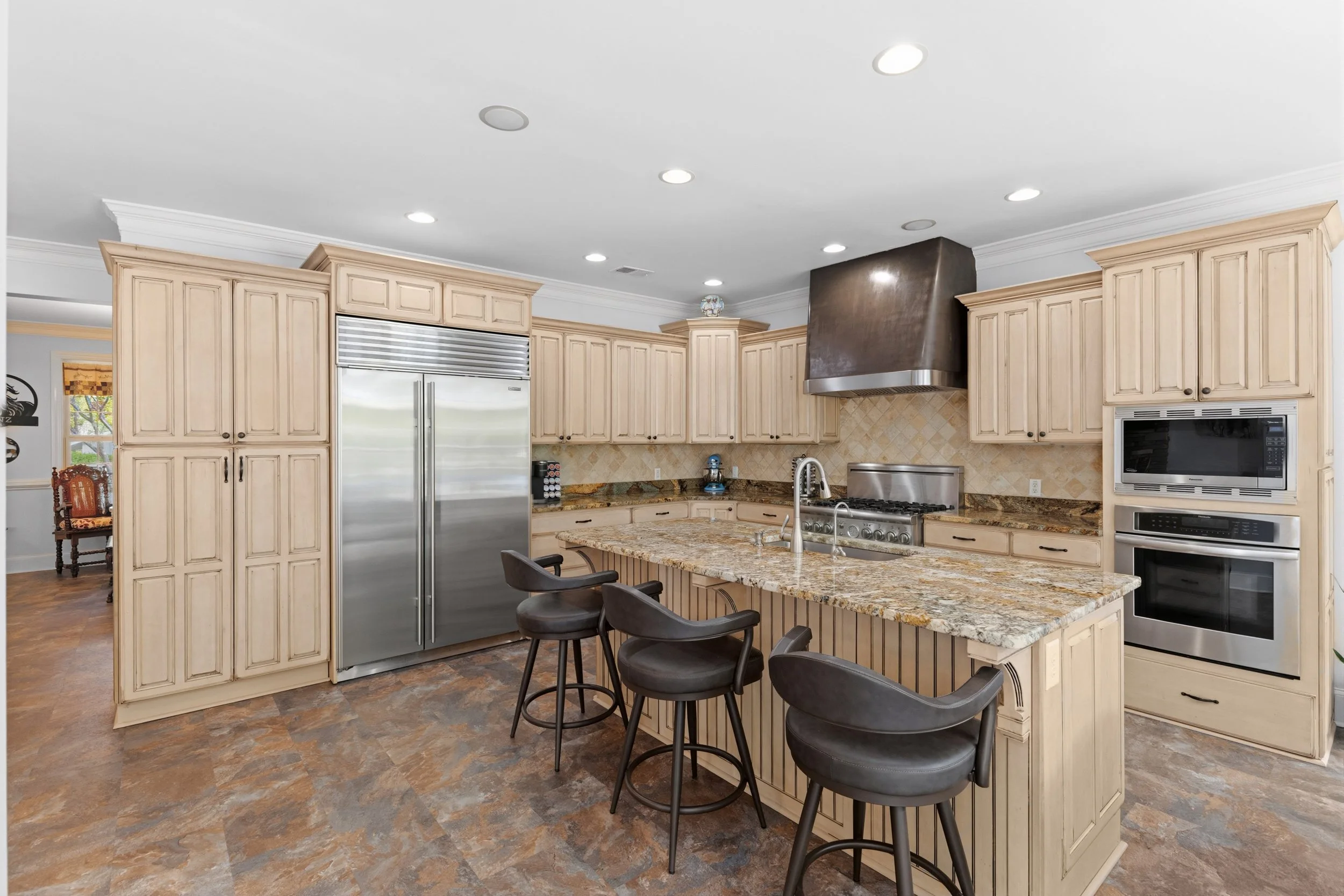 Kitchen with beige cabinets, stainless steel refrigerator and oven, granite island with four black bar stools, microwave, stovetop, and tiled backsplash.
