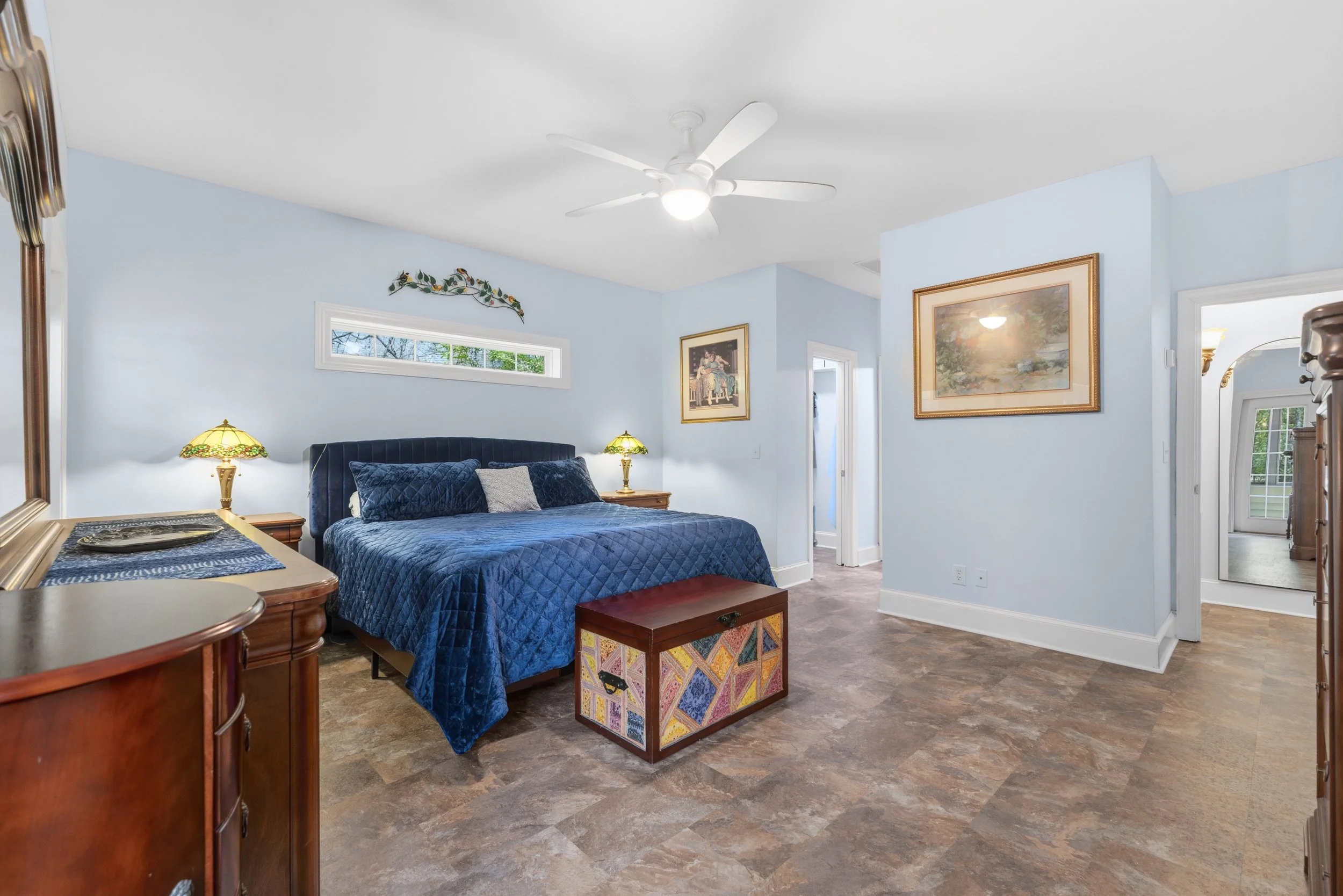 Bedroom with light blue walls, a large bed with dark blue quilt and pillows, two wooden nightstands with lamps, a wooden dresser with a mirror, framed artwork on the walls, a small horizontal window, ceiling fan, and a colorful wooden storage chest i
