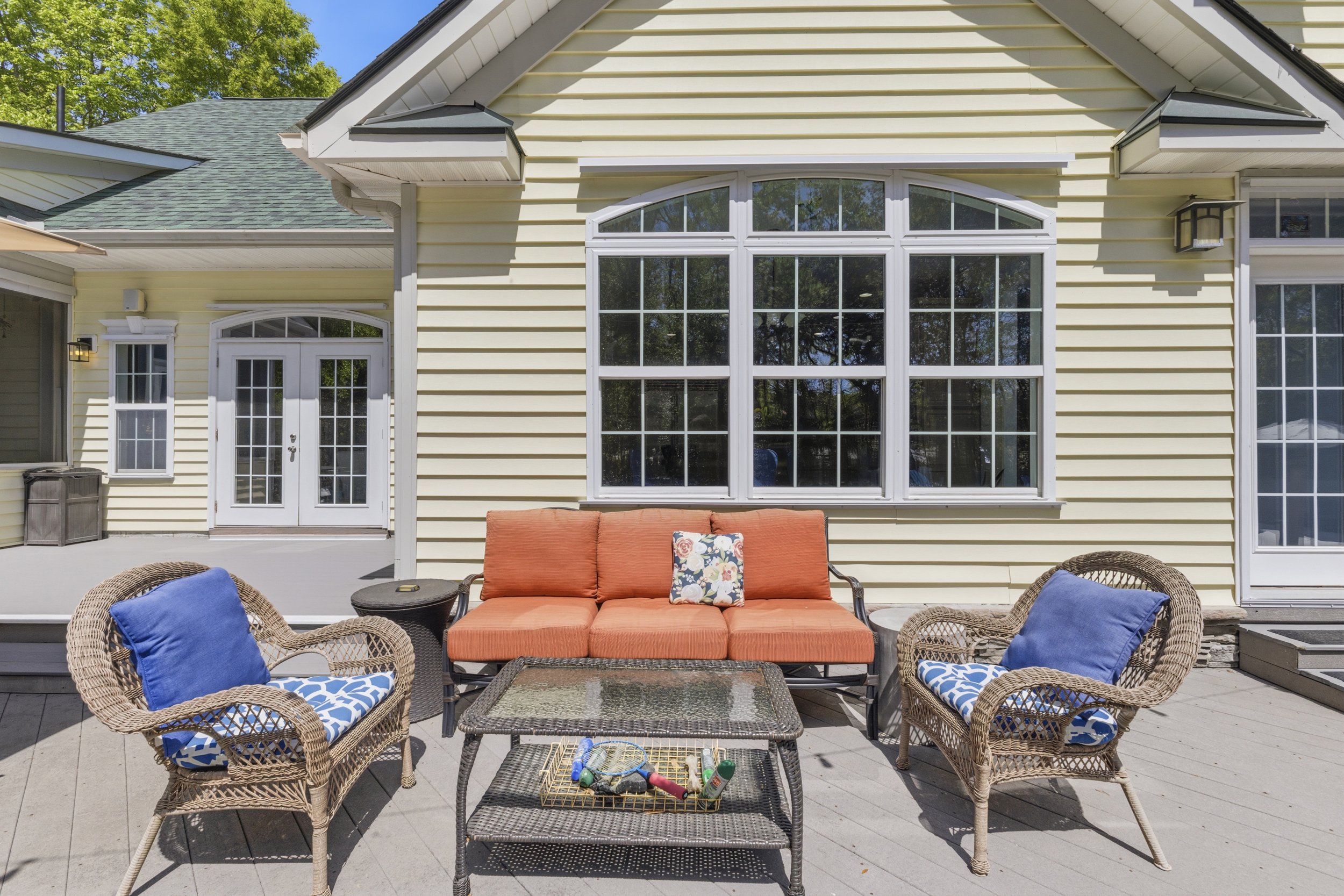 Outdoor patio with wicker chairs, orange cushioned sofa, and a glass-top table in front of a yellow house with large windows and French doors, surrounded by greenery.