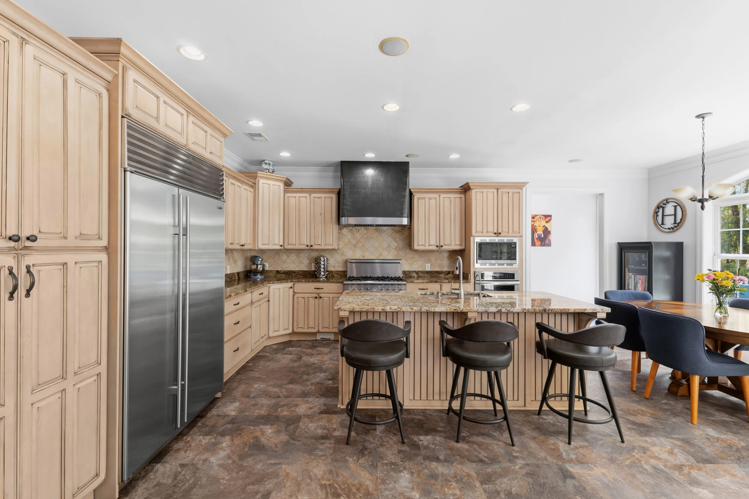 Kitchen with beige cabinets, stainless steel fridge, gas stove with black vent hood, granite countertops, island with three black stools, and a dining area with a table, blue chairs, and a flower vase near a window.