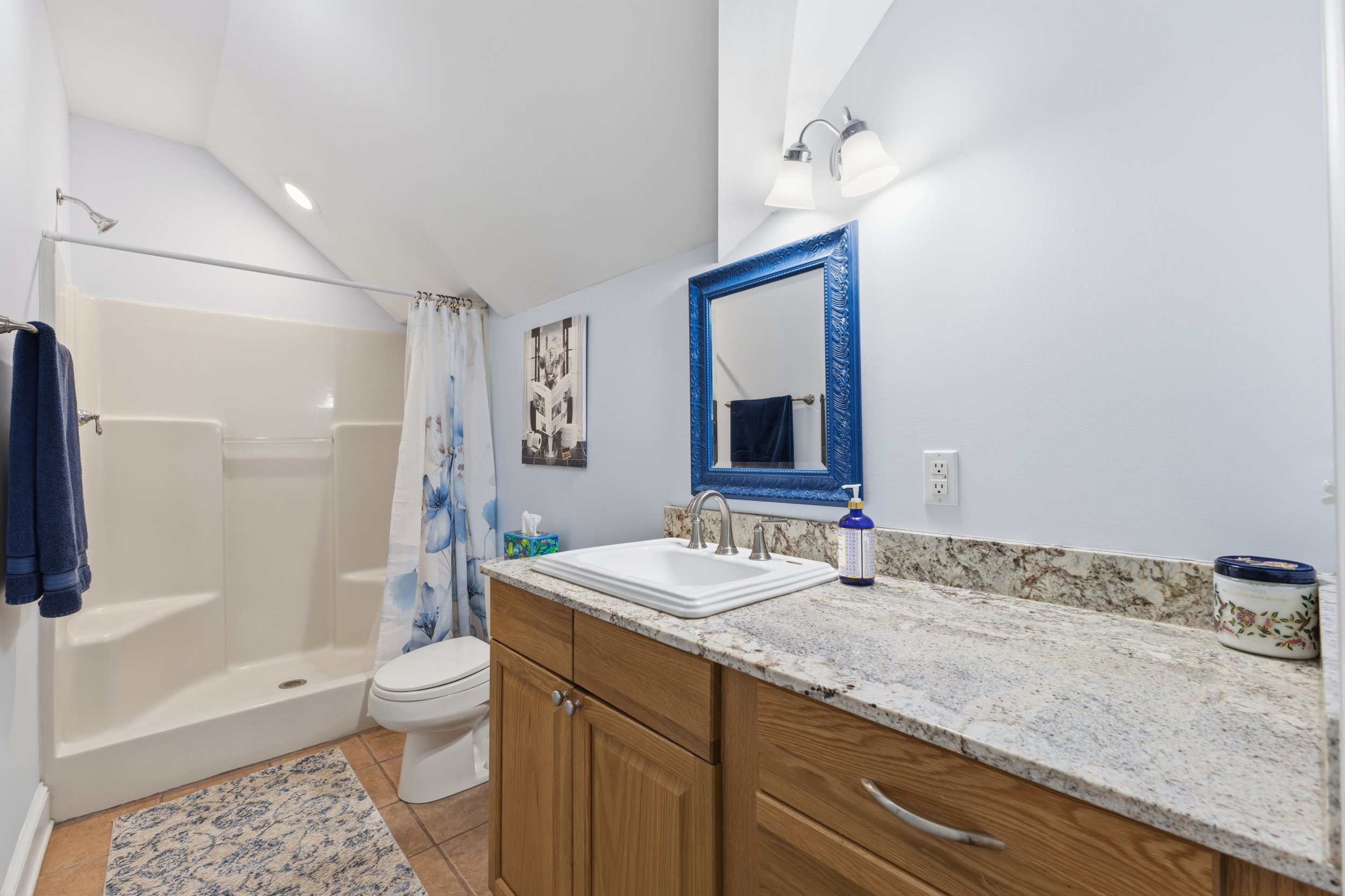 Bathroom with a white shower, toilet, wooden vanity with granite countertop, blue framed mirror, and decorative items.