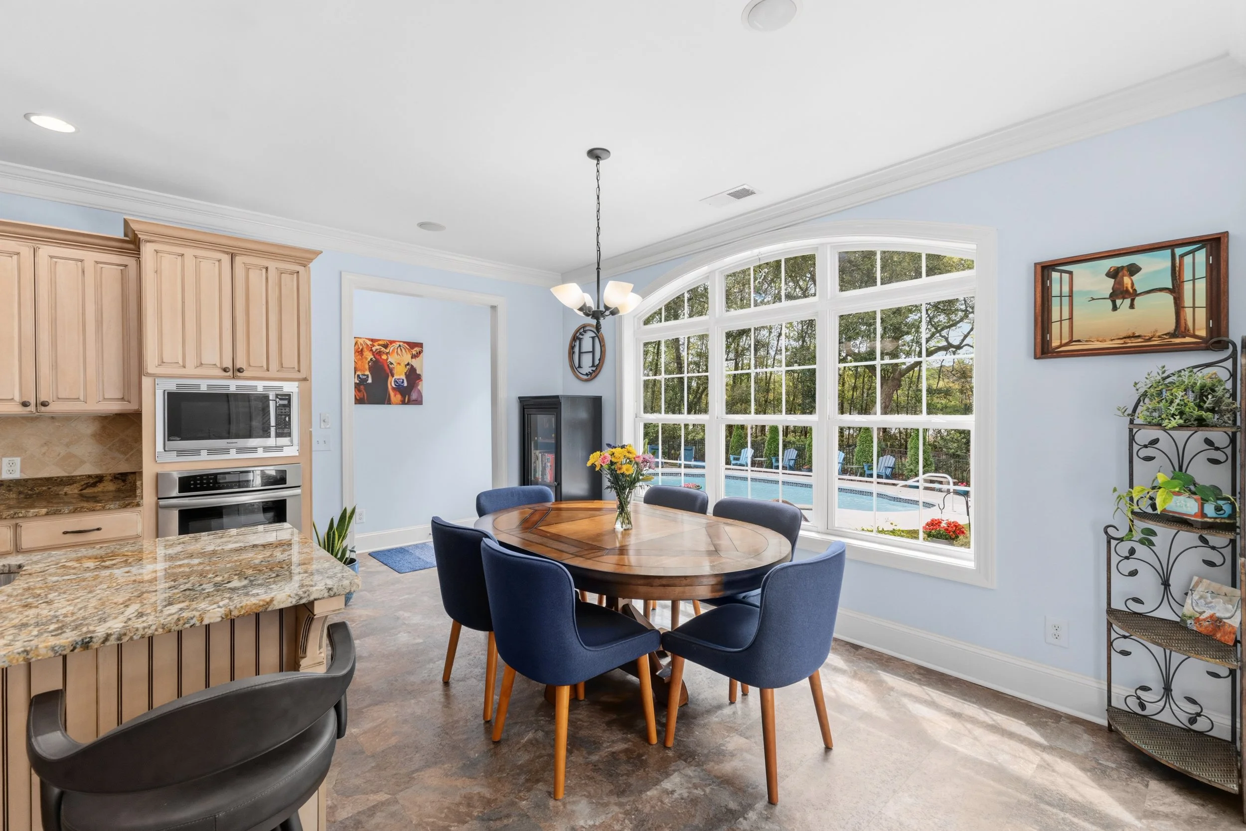 Bright kitchen and dining area with large bay window, round wooden dining table with six navy chairs, kitchen with wood cabinets, granite countertop, and built-in microwave and oven, view of pool outside