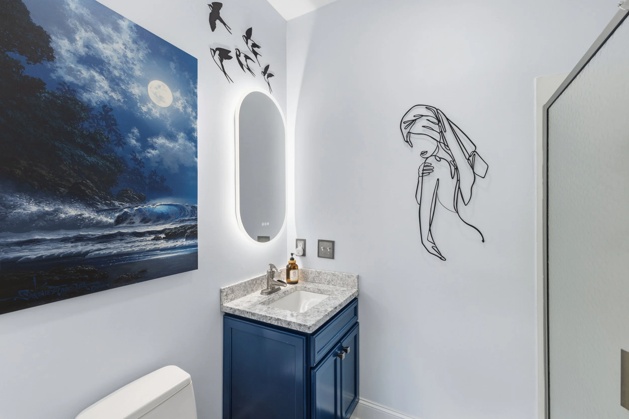 A small bathroom with a white wall featuring a large ocean landscape painting, a decorative wire art of a woman, a mirror with backlit LED, a blue cabinet with a granite countertop, a sink, and a soap dispenser.