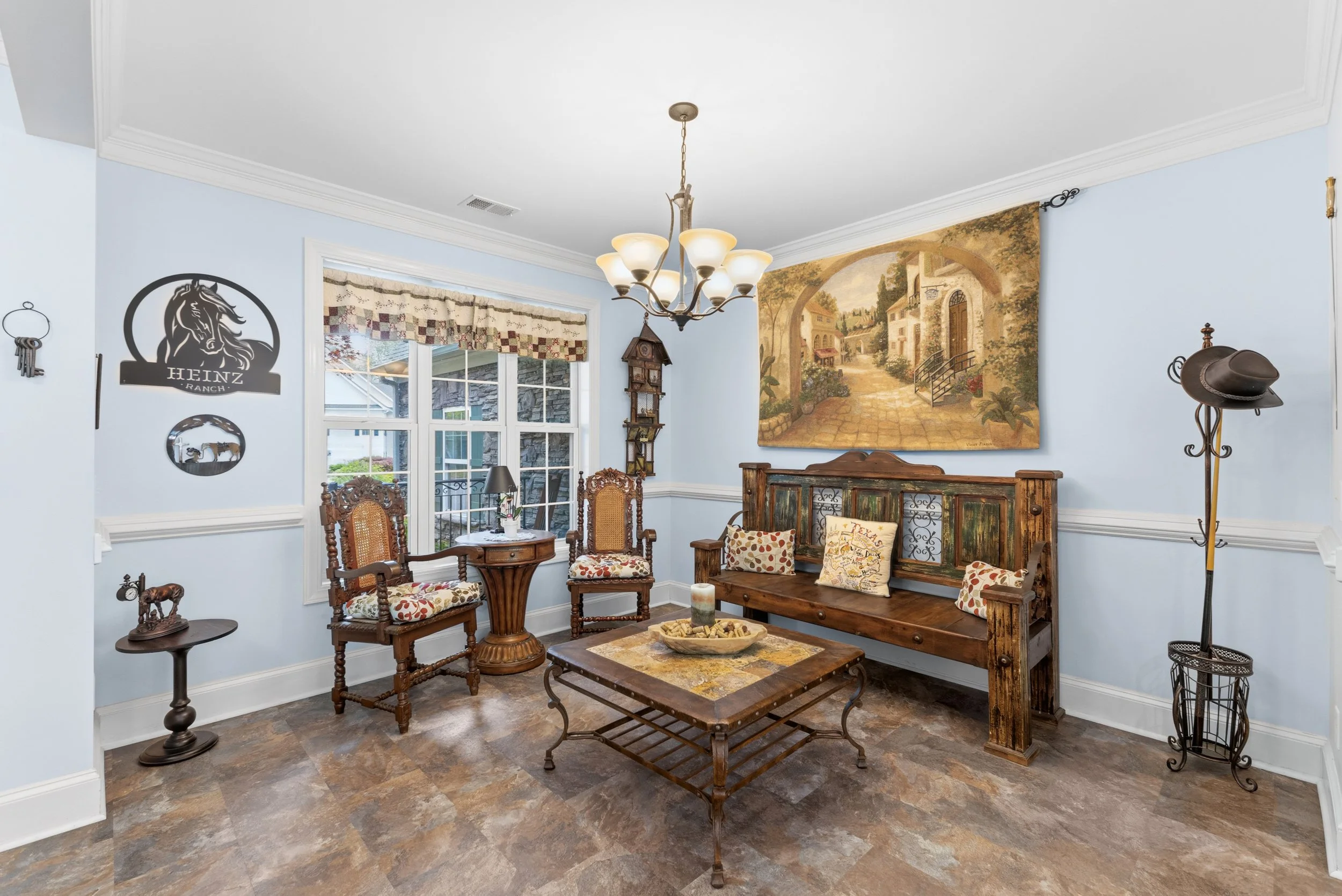 Living room with light blue walls, vintage wooden furniture, a chandelier, a large mural of a European street scene, and a window with a valance.