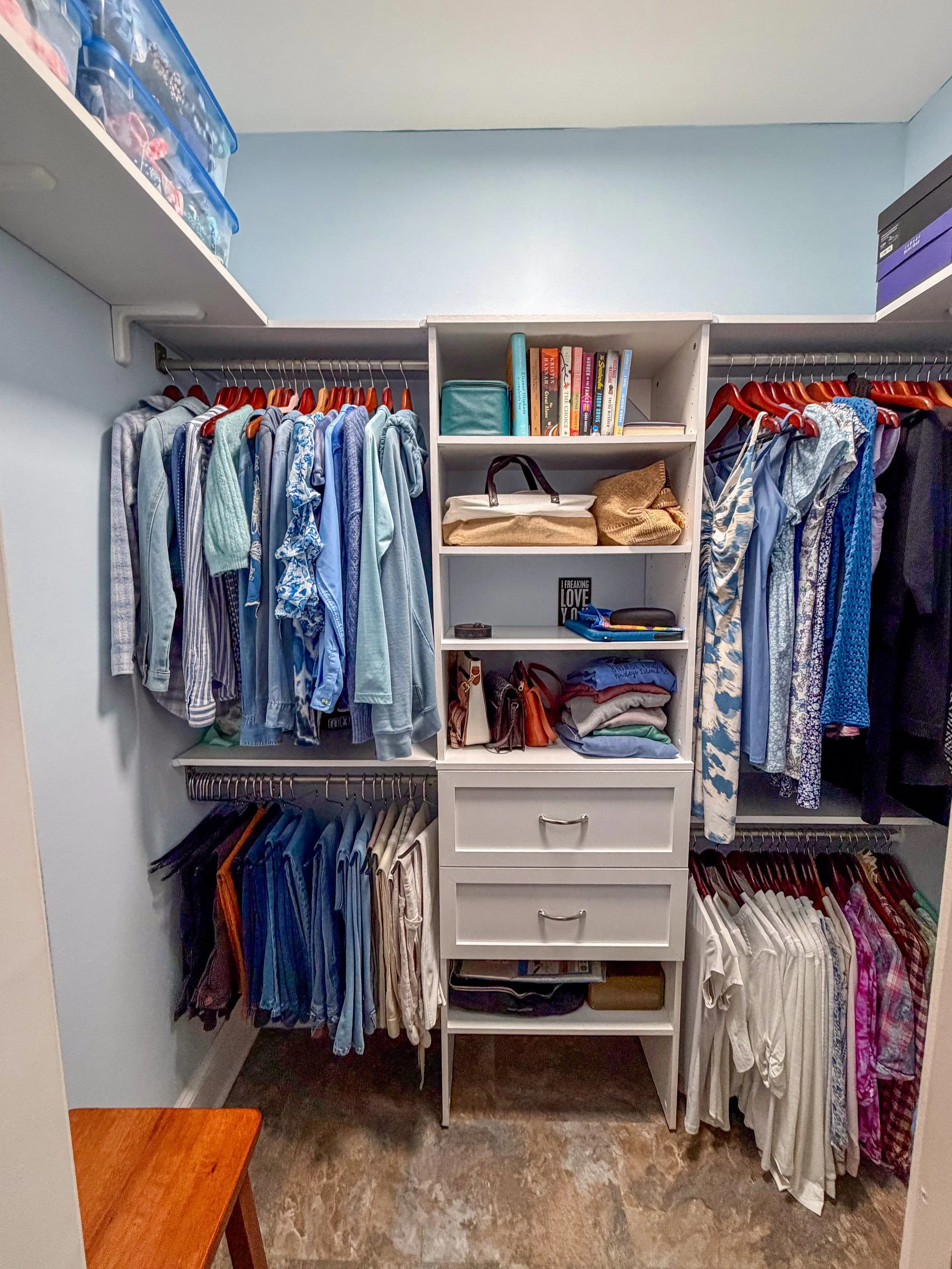 A closet with clothes hanging on rods, including shirts and pants, and a white shelving unit with books, bags, and folded clothes.
