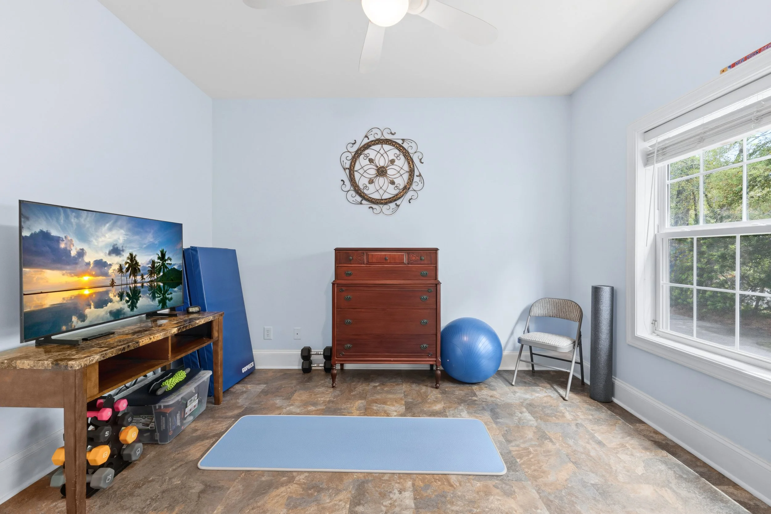 A home gym room with light blue walls, a ceiling fan, a large window letting in natural light, a wooden dresser, a blue exercise ball, a gray chair, a foam roller, dumbbells, a yoga mat, and a flat-screen TV showing a tropical beach scene.