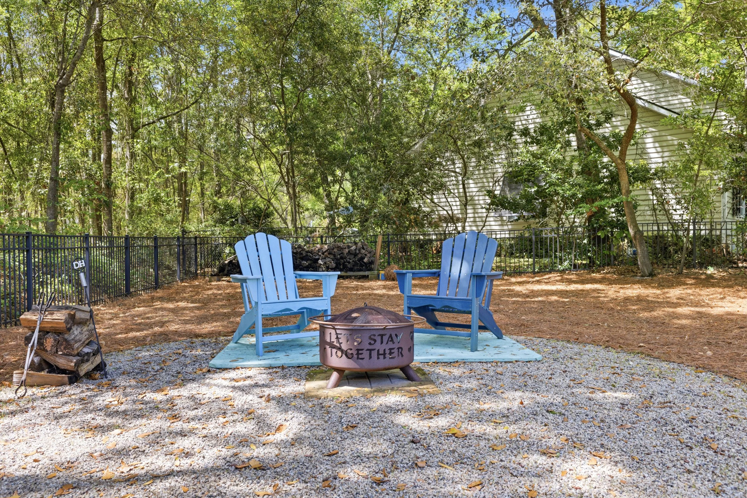 A backyard patio area with two blue Adirondack chairs facing each other over a fire pit that has the words "Let's Stay Together" written on it. The area is enclosed by a black metal fence, with trees and a house in the background. There's a pile of f
