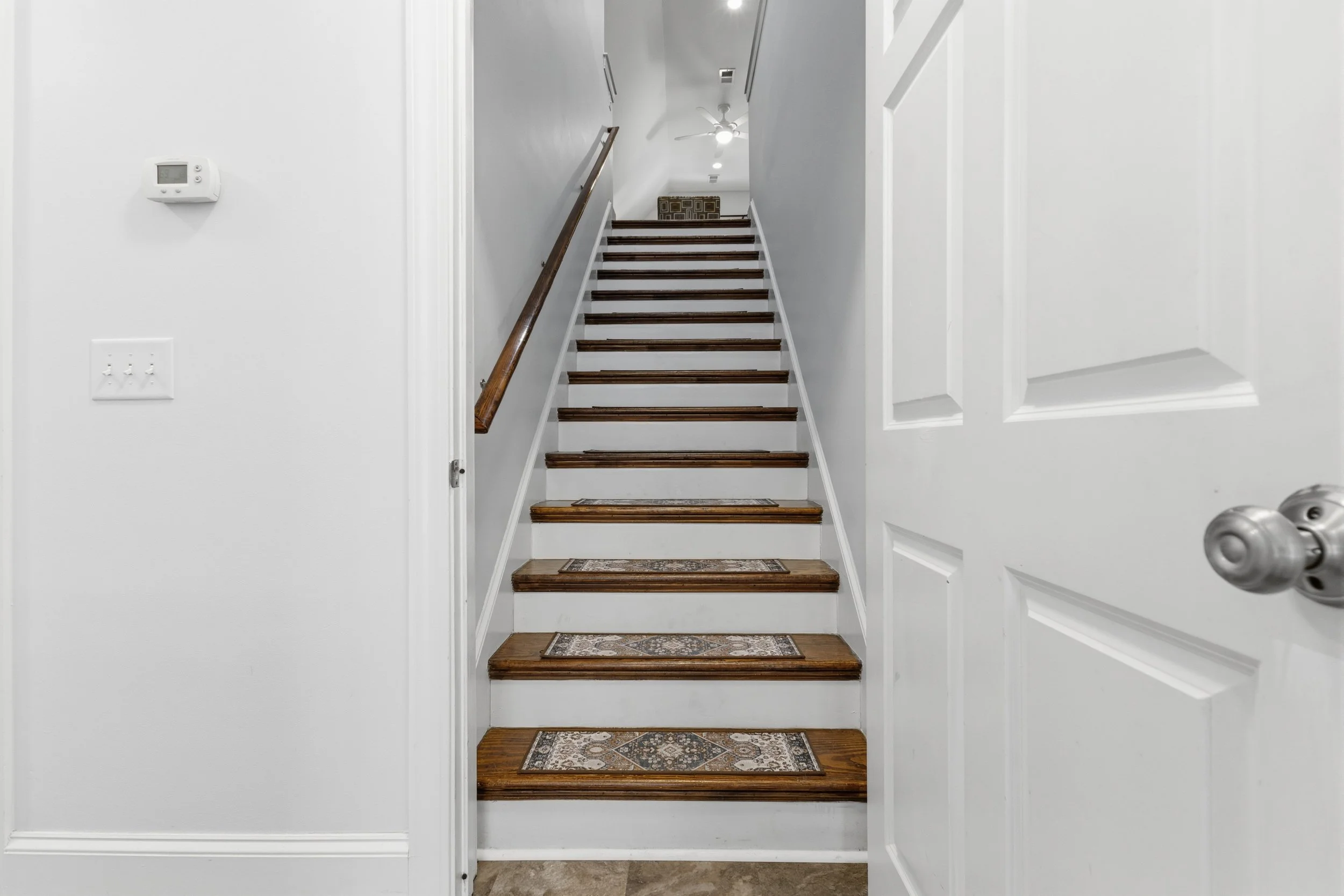 Indoor staircase with wooden steps and white risers, a dark wooden handrail on the left, a white wall on the right, and a ceiling fan at the top.