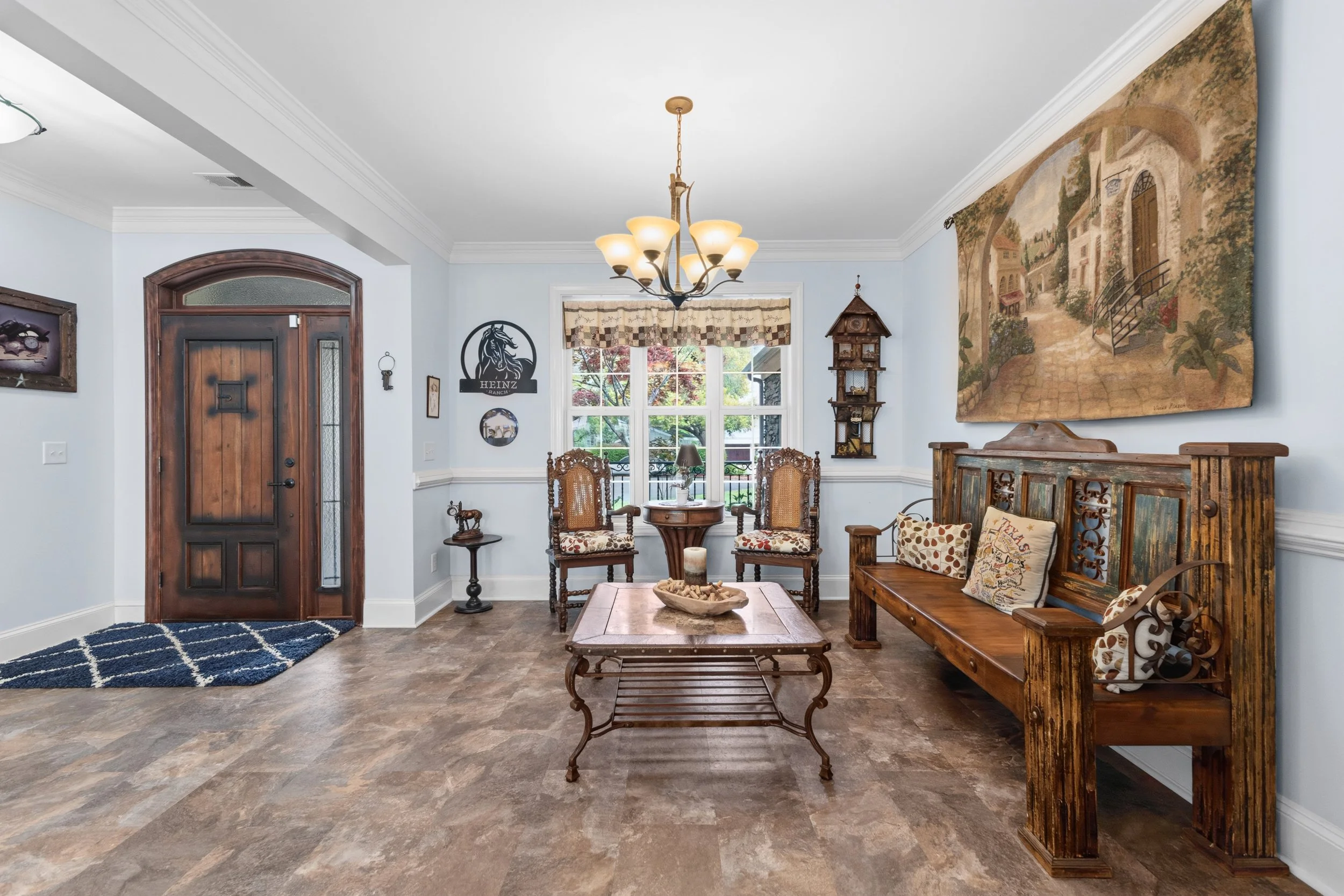 Living room with wooden front door, two armchairs, a wooden bench with cushions, a wooden coffee table, a chandelier, a window with plaid curtains, and wall decorations including a large landscape painting and a cuckoo clock.
