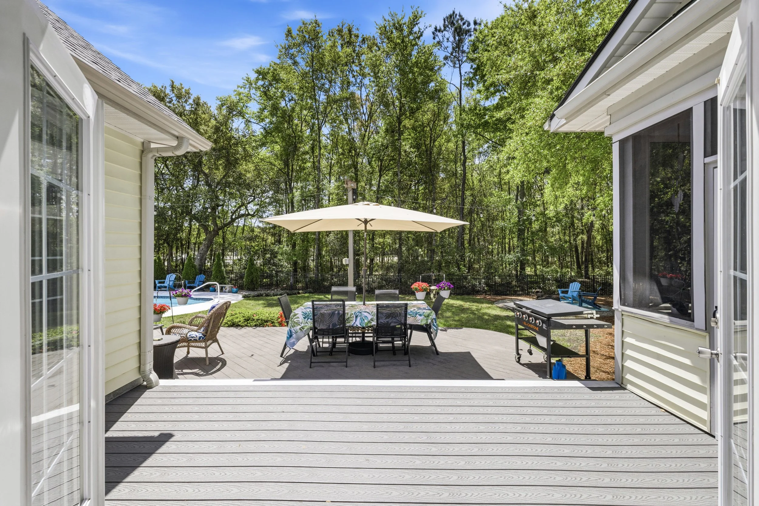 View from a house patio looking out onto a backyard with a table and chairs under an umbrella, a grill, and a garden with trees and outdoor furniture.