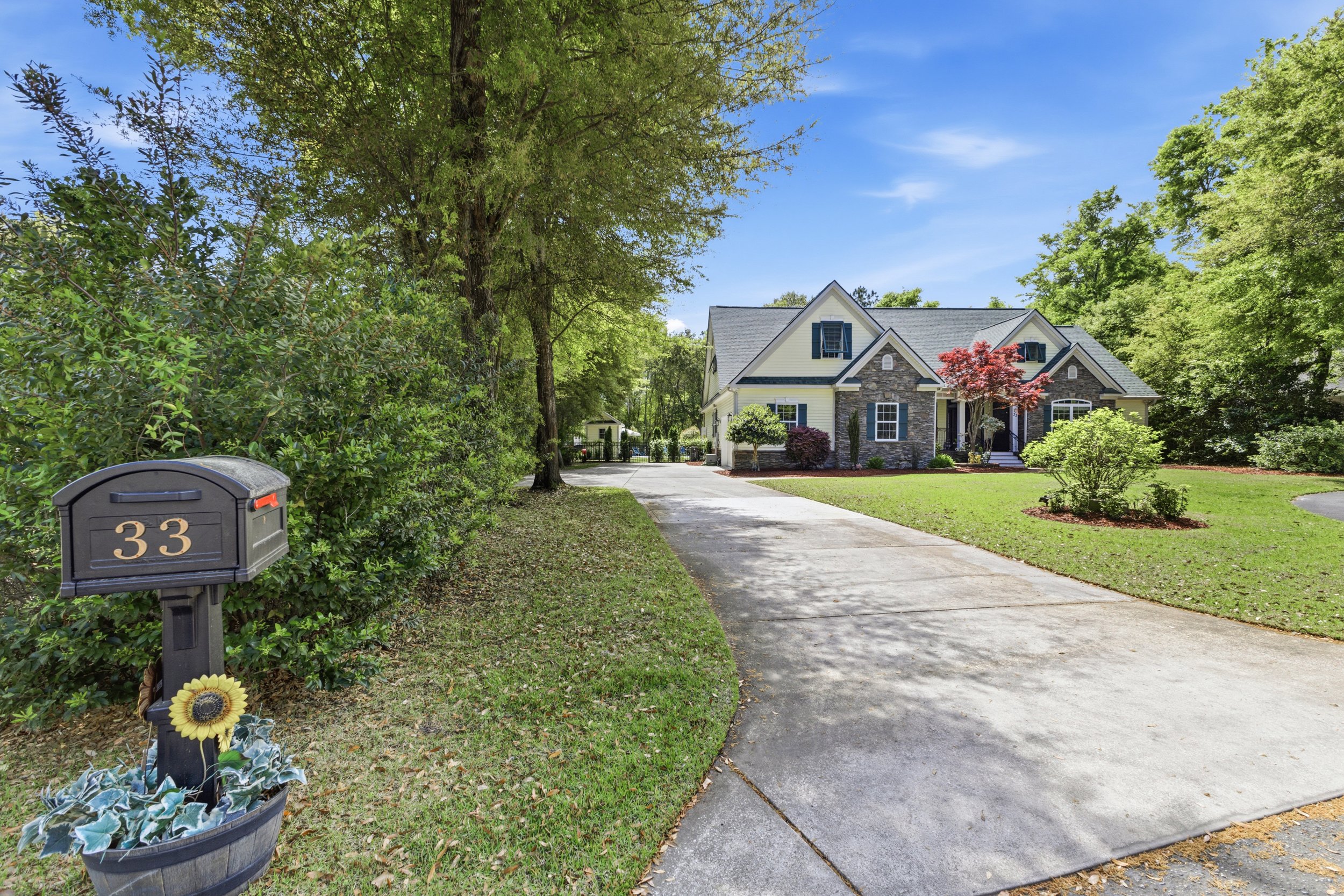 A suburban house with a stone and siding exterior, surrounded by green lawn and mature trees, with a concrete driveway leading up to it, and a mailbox with the number 33 in the foreground.