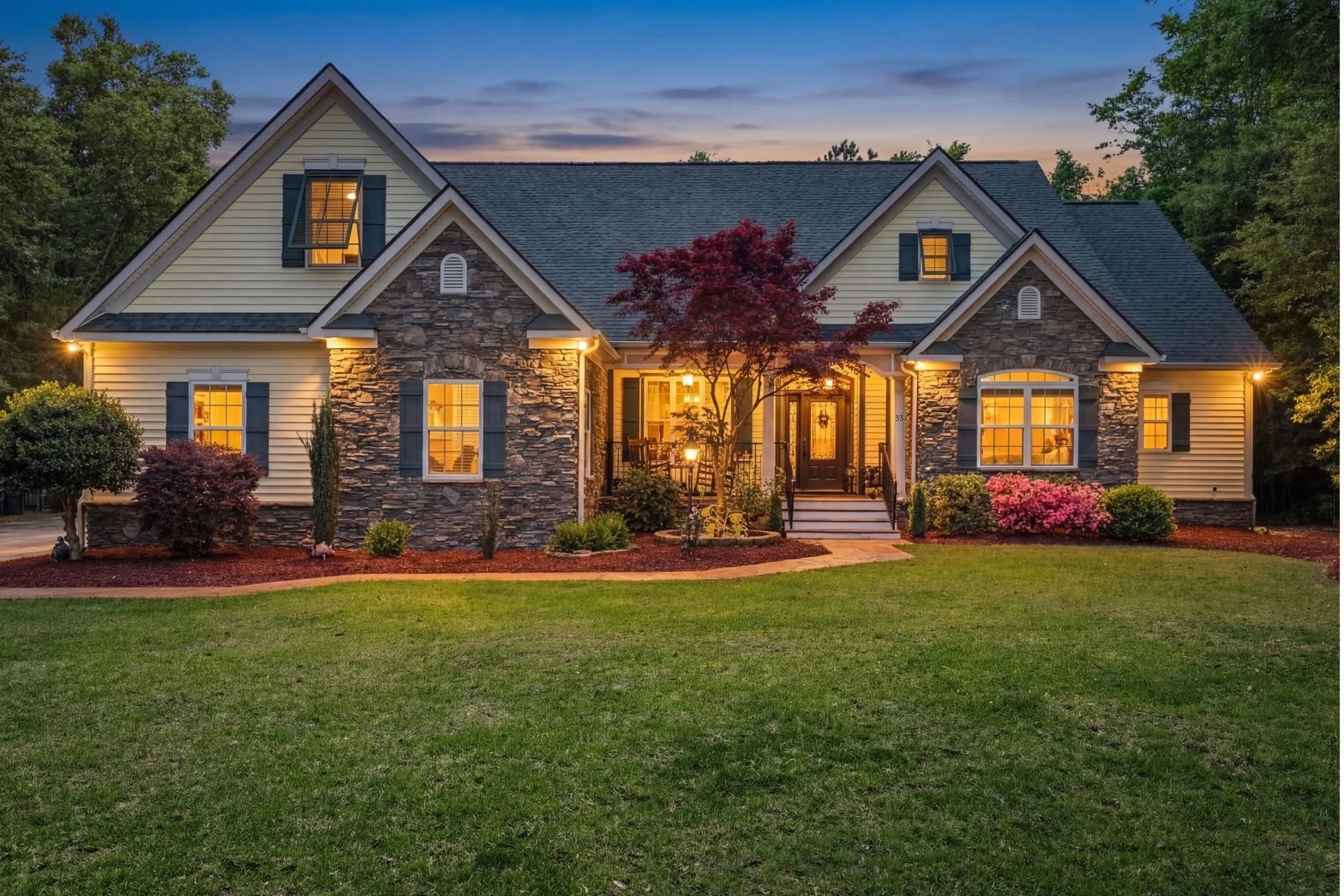 A two-story house with stone and beige siding exterior, lit up at dusk, surrounded by a well-maintained lawn and landscaped bushes and trees.