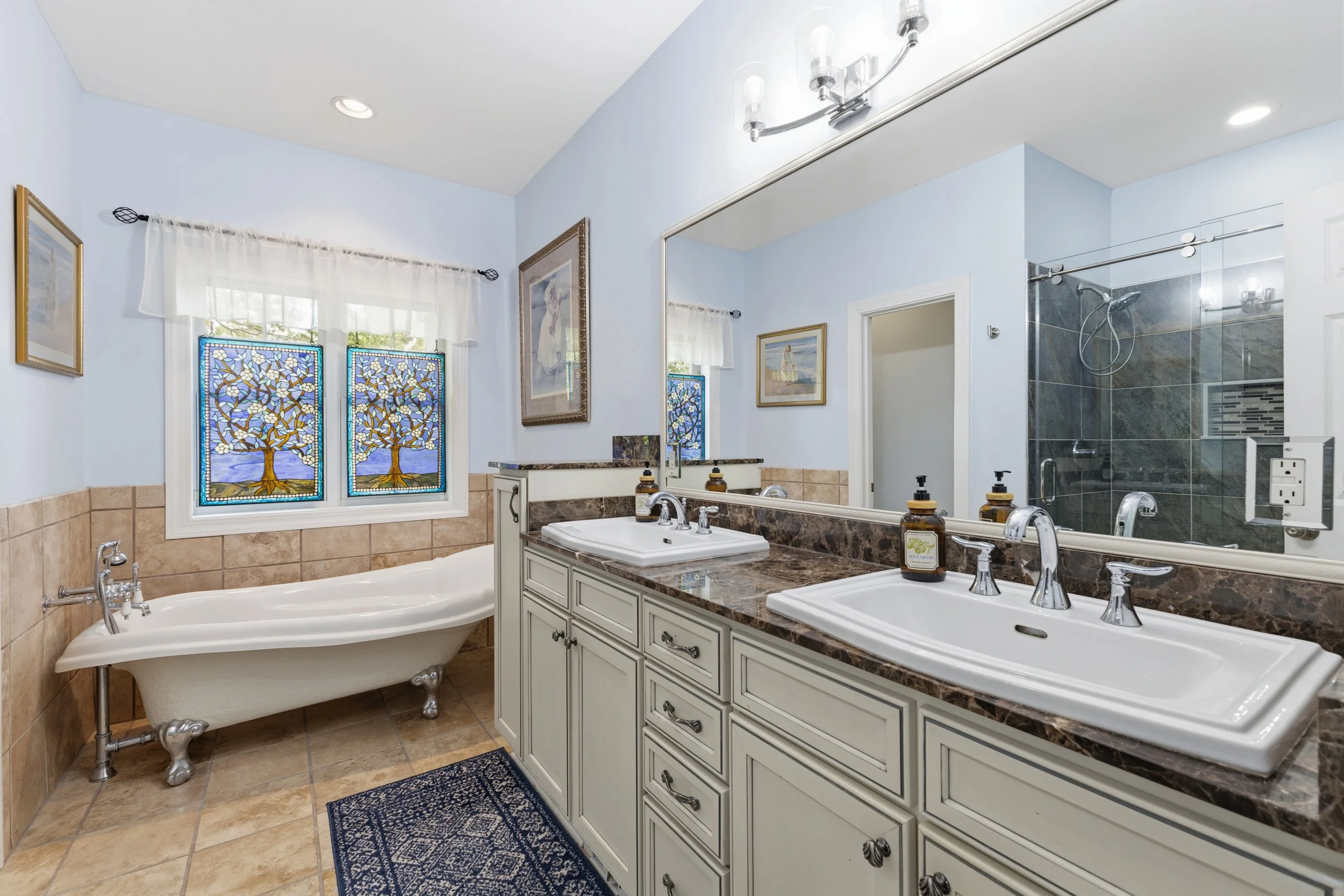 Bathroom with double sink vanity, large mirror, bathtub, and walk-in shower, decorated with stained glass window, framed artwork, and a blue rug.