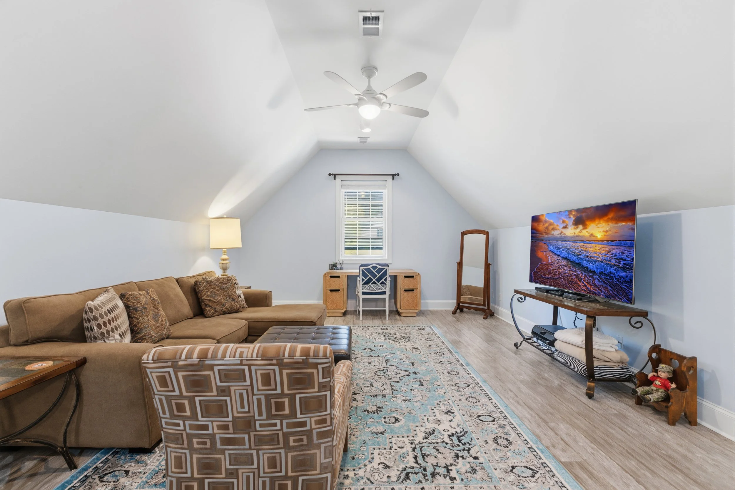 Living room with sloped ceiling, beige sectional sofa, patterned armchair, area rug, small desk with chair beneath window, standing mirror, flat-screen TV on a stand, and corner with decorative items.