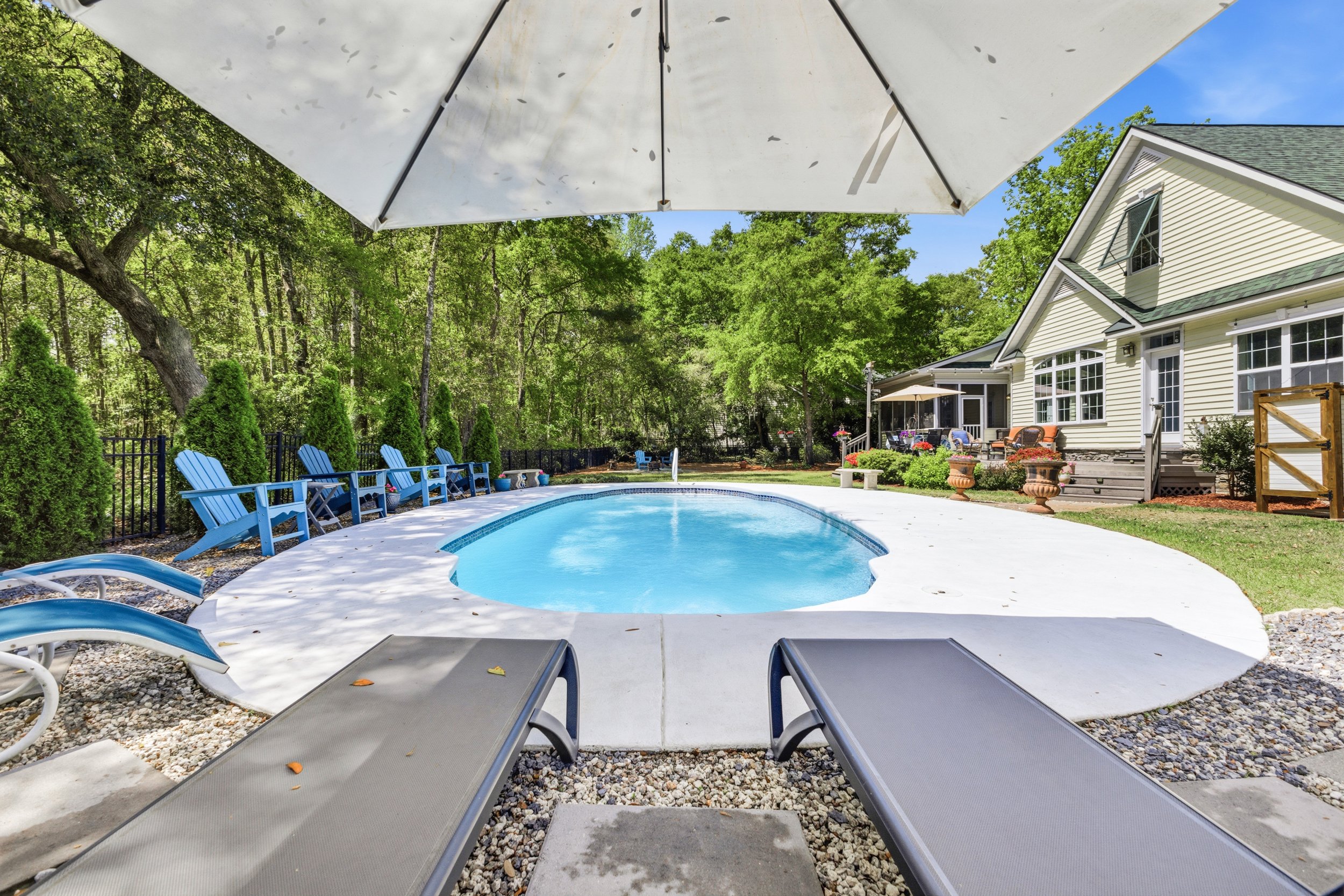 Residential backyard with pool, lounge chairs, umbrellas, and lush green trees.