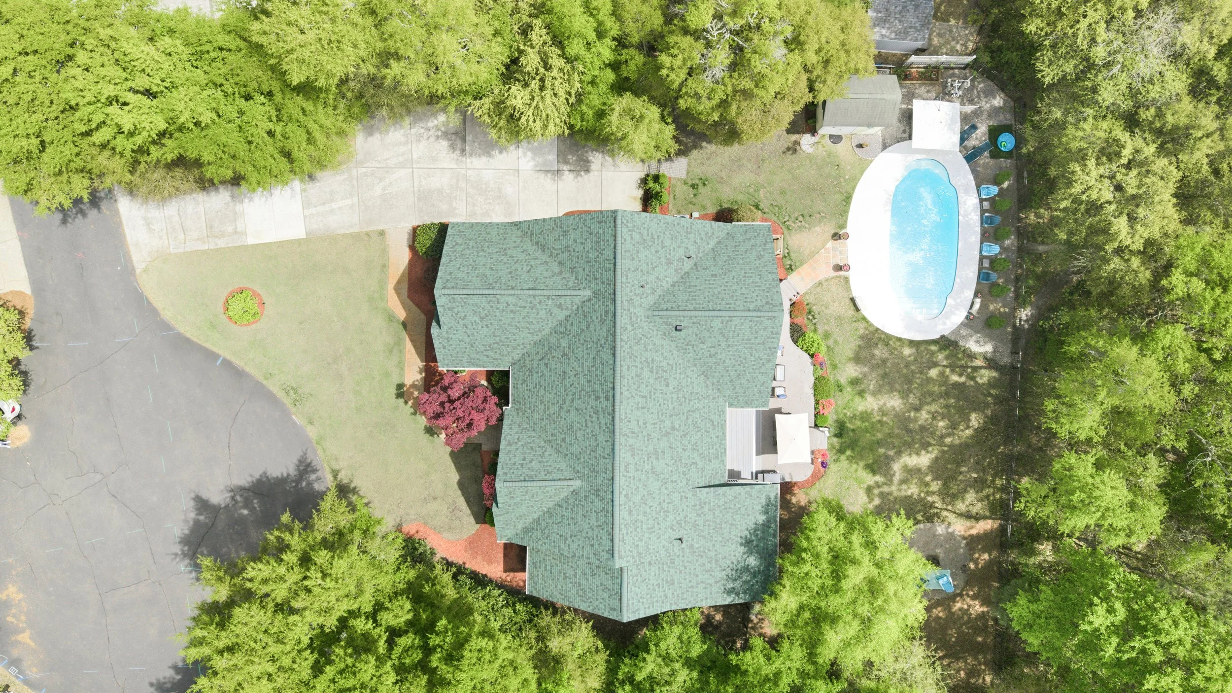 An aerial view of a house with a green roof, surrounded by trees, a driveway, a shaded lawn, and an above-ground swimming pool with a concrete patio and chairs.