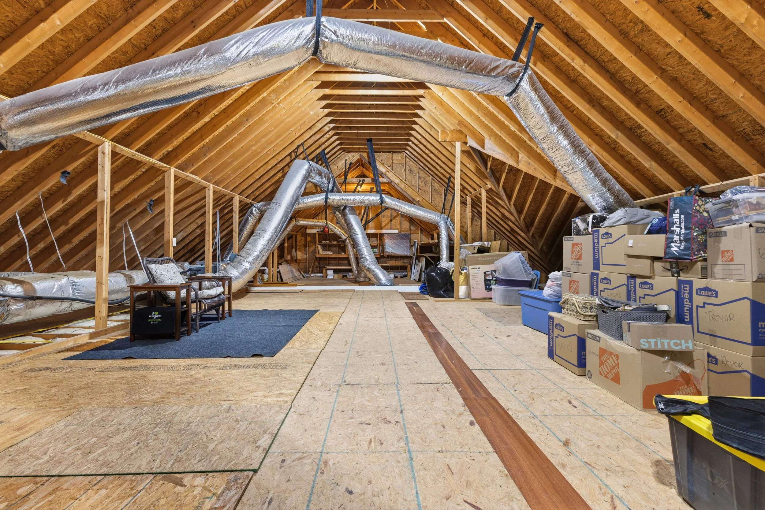 Attic space with exposed wooden beams, ducts, and insulation; stacked boxes and miscellaneous items on the right side, with chairs and rugs on the left.