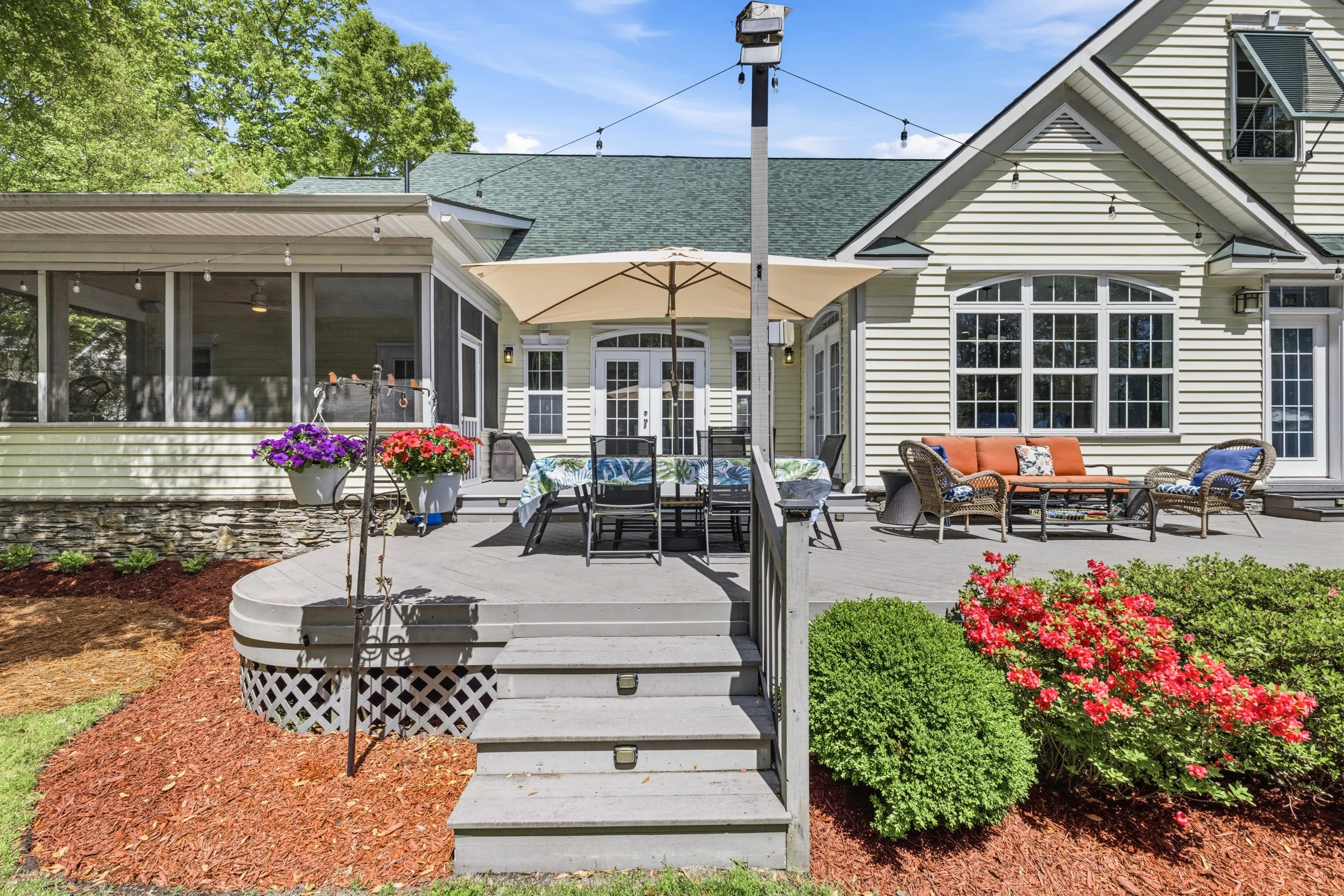 Backyard patio with outdoor furniture, a table with an umbrella, potted flowers, and decorative bushes, in front of a house with large windows and a screened porch on a sunny day.