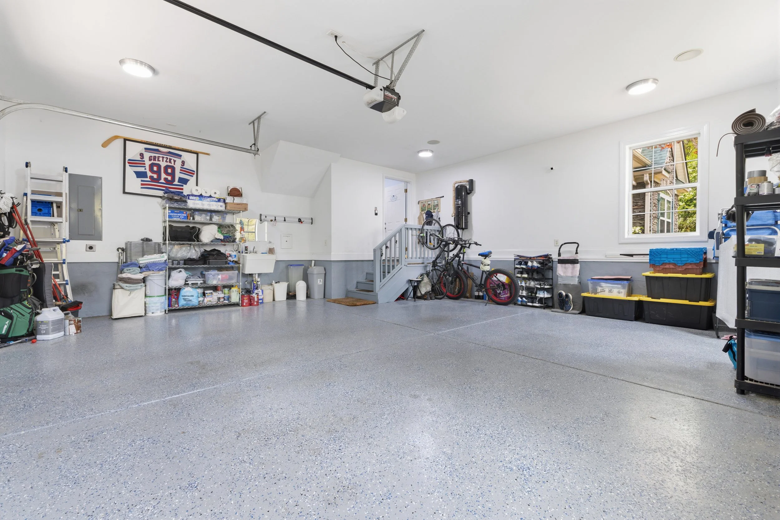 Empty garage with shelves, storage bins, bicycles, and garage door opener, with a window and staircase.