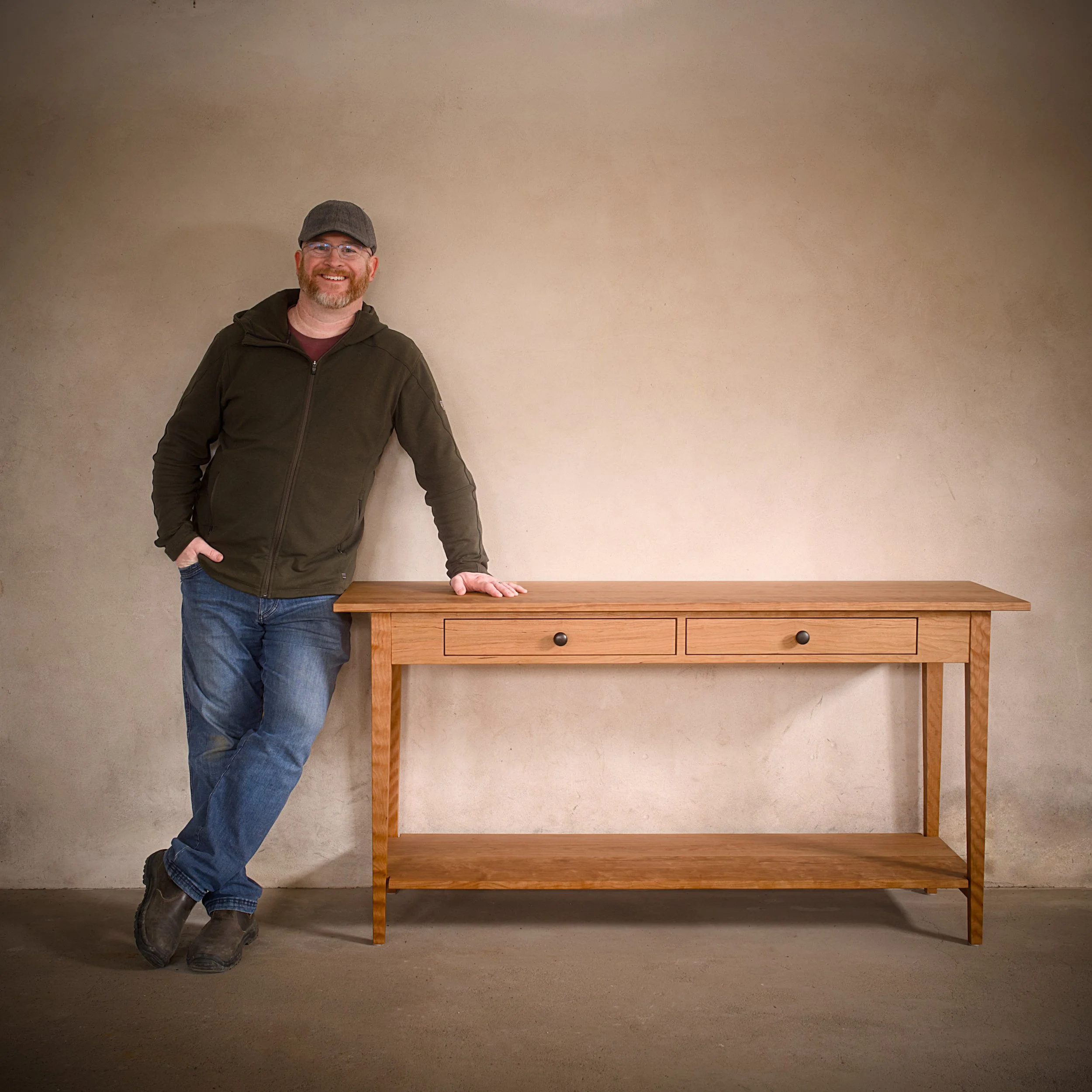 A man standing next to a wooden Shaker-style console table with two drawers against a beige wall.
